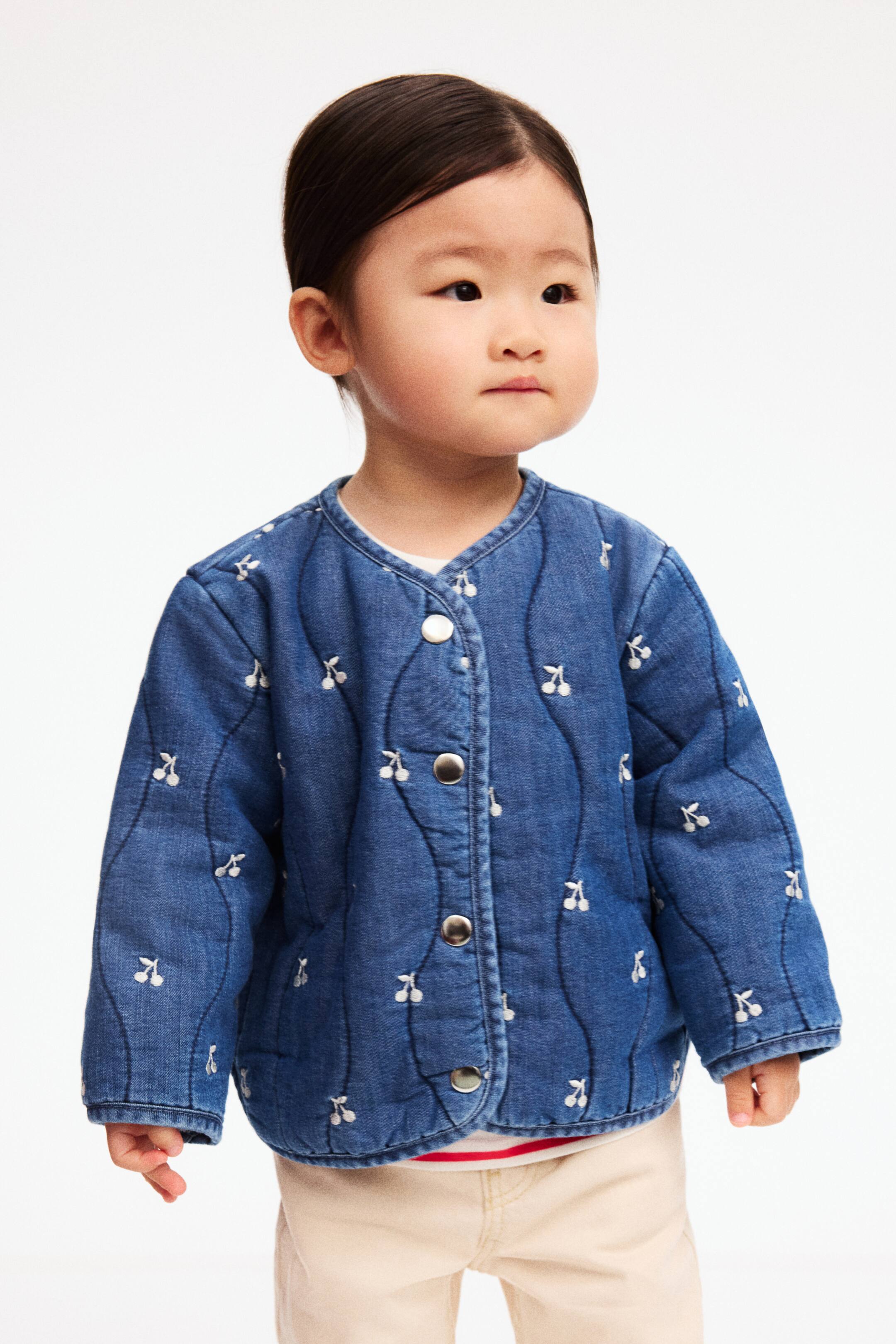 Ampliar la imagen: Se presenta una chaqueta acolchada de aspecto mezclilla azul, vista de frente y ligeramente inclinada hacia la derecha, con pespuntes ondulados y pequeñas cerezas bordadas en blanco. Se cierra al frente con botones a presión en tono plateado, presenta un cuello sin solapa y está superpuesta a una camiseta interior blanca cuyo dobladillo a rayas queda visible debajo.