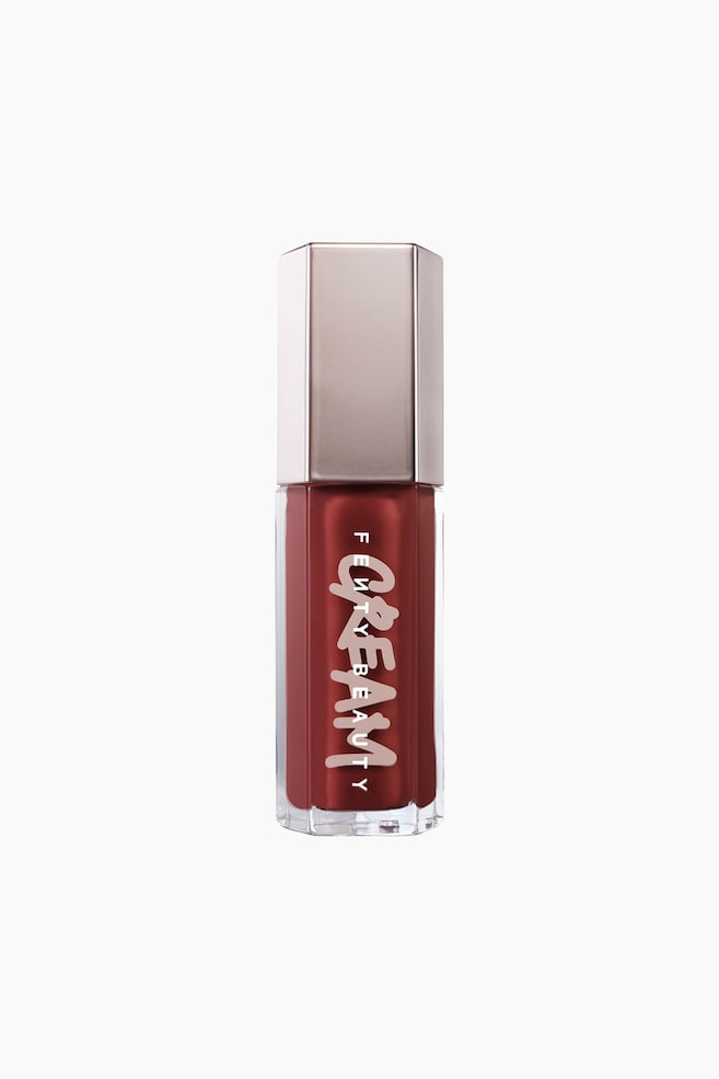 En Fenty Beauty-lipgloss-flaske stÃ¥r oprejst og fremviser et sekskantet, gennemsigtigt rÃ¸r fyldt med en dyb rÃ¸dbrun cream gloss. Forsiden af rÃ¸ret har ordet "CREAM" i en stiliseret, fed lyserÃ¸d skrifttype trykt lodret over "FENTY BEAUTY" i hvidt, afsluttet med et metallisk sÃ¸lvfarvet sekskantet lÃ¥g. - 1