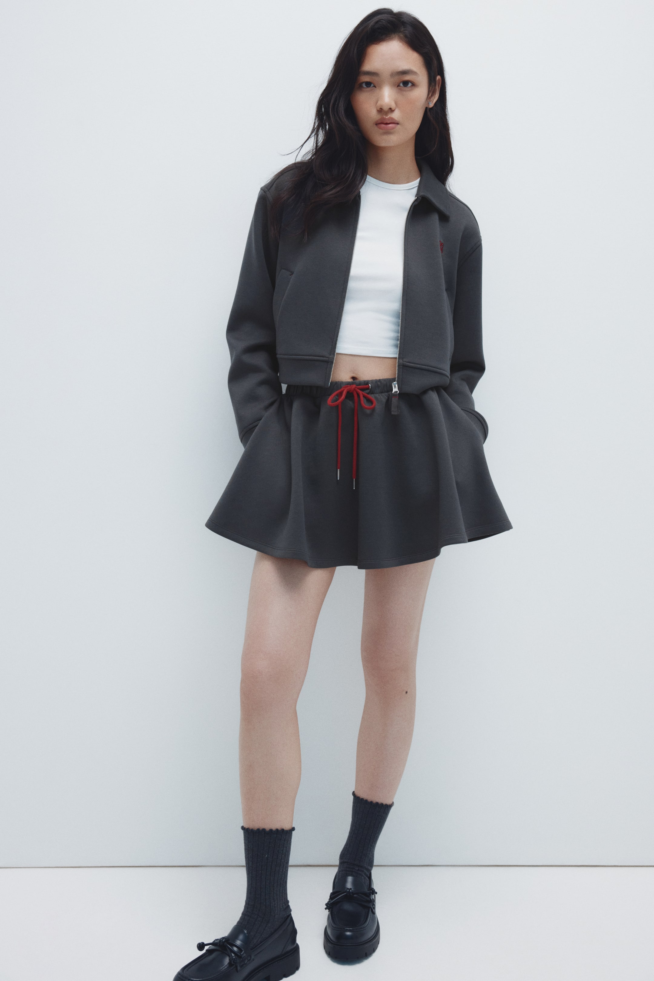 拡大画像を表示: A dark grey cropped jacket and matching flared mini skirt are being presented from a front view.ジャケットはフルジップの前面、カラー、左胸に小さな赤いエンブレムが特徴で、白いクロップトップの上に重ねられています。スカートは、赤い目立つドローストリングが付いた伸縮性のあるウエストバンドを特徴としています。
