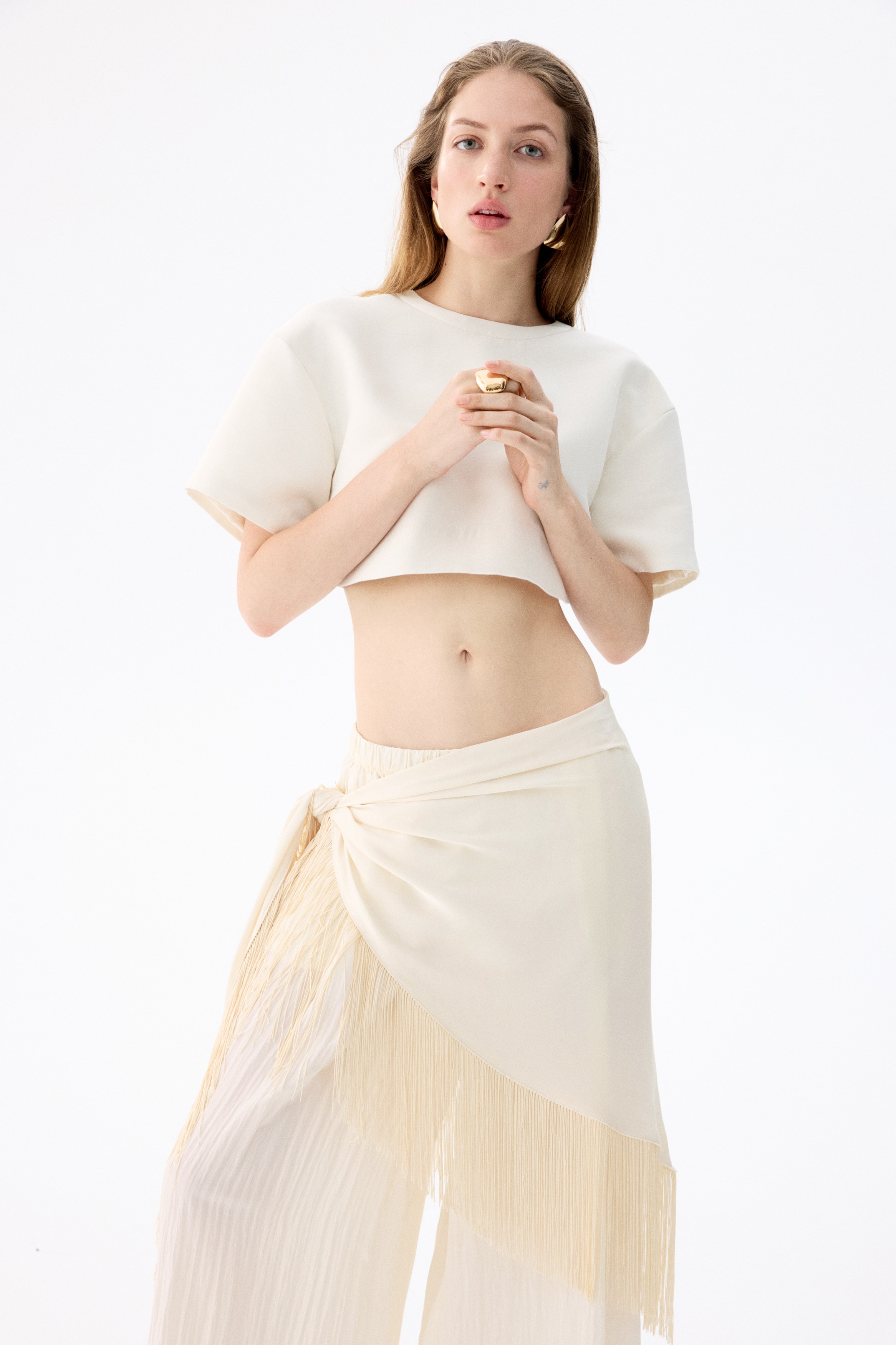 A cream-coloured fringe wrap skirt showcasing a draped front design. Long fringes extend along the asymmetrical bottom edge, cascading from a prominent knot on the right side.