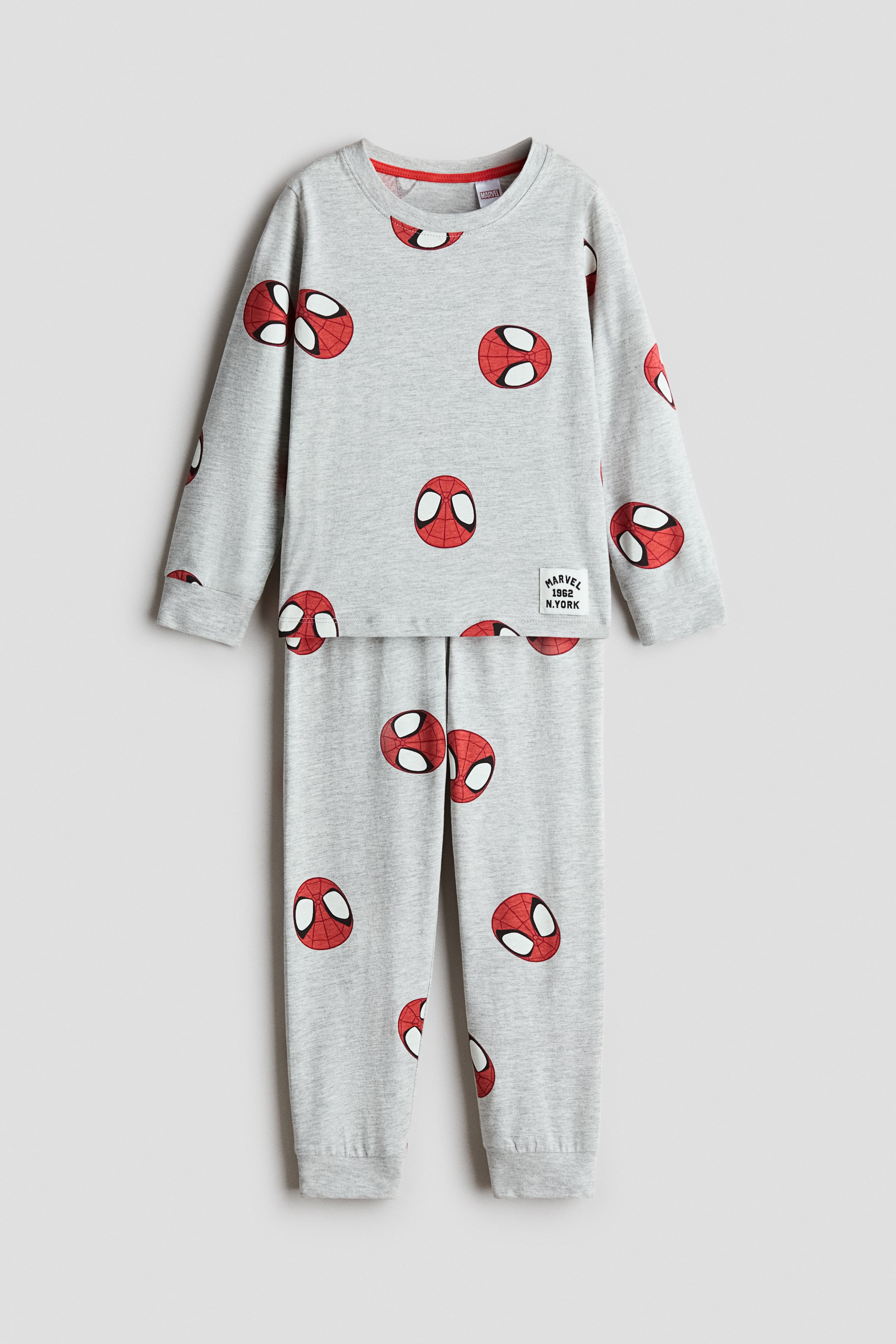View larger image: A two-piece pyjama set in light grey melange is shown from the front, featuring an all-over print of red and black Spider-Man masks. The long-sleeved top shows a round ribbed neckline, ribbed cuffs, and a small white graphic patch near the hem, while the matching pants feature ribbed hems.