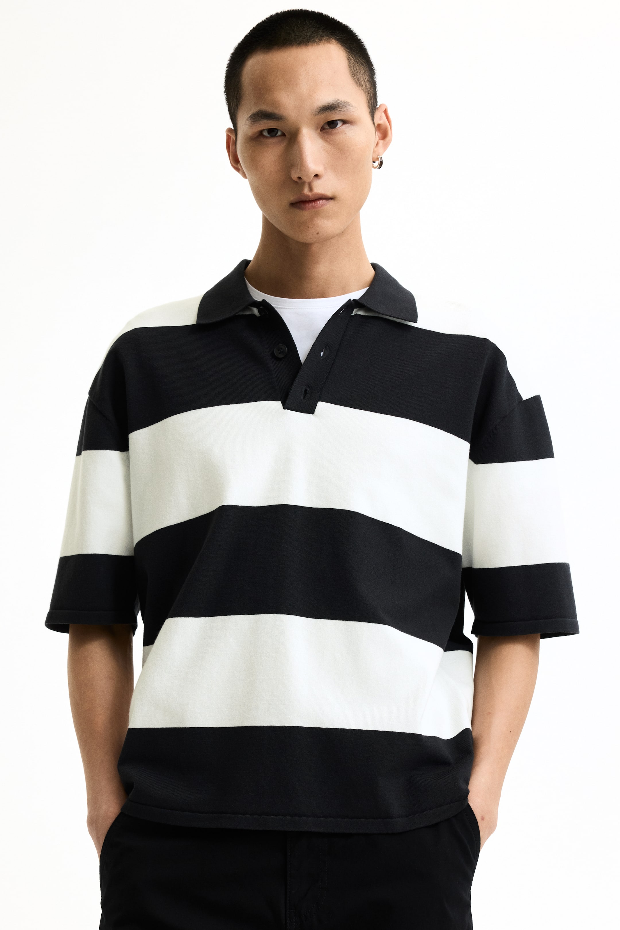 View larger image: A black and white striped polo shirt featuring broad horizontal stripes, a pointed black collar, and a three-button placket. It displays short, wide sleeves and a heavyweight knit texture, with a white crew-neck t-shirt visible underneath.