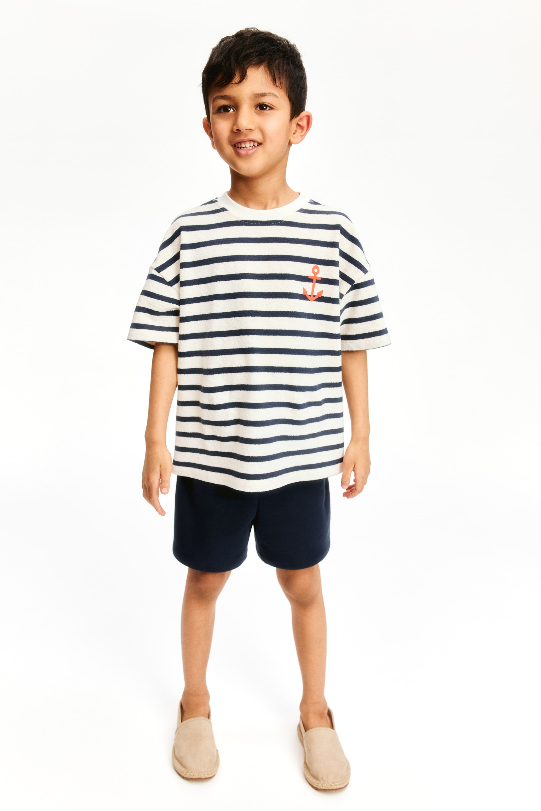 Grösseres Bild ansehen: Ein Kinderoutfit wird präsentiert, bestehend aus einem creme- und marineblau gestreiften Frottee-T-Shirt mit weiten kurzen Ärmeln und einem orangefarbenen Ankeremblem auf der linken Brust. Marineblaue Shorts und beigefarbene Espadrille-Slipper vervollständigen das Ensemble, alle nach vorne gerichtet.