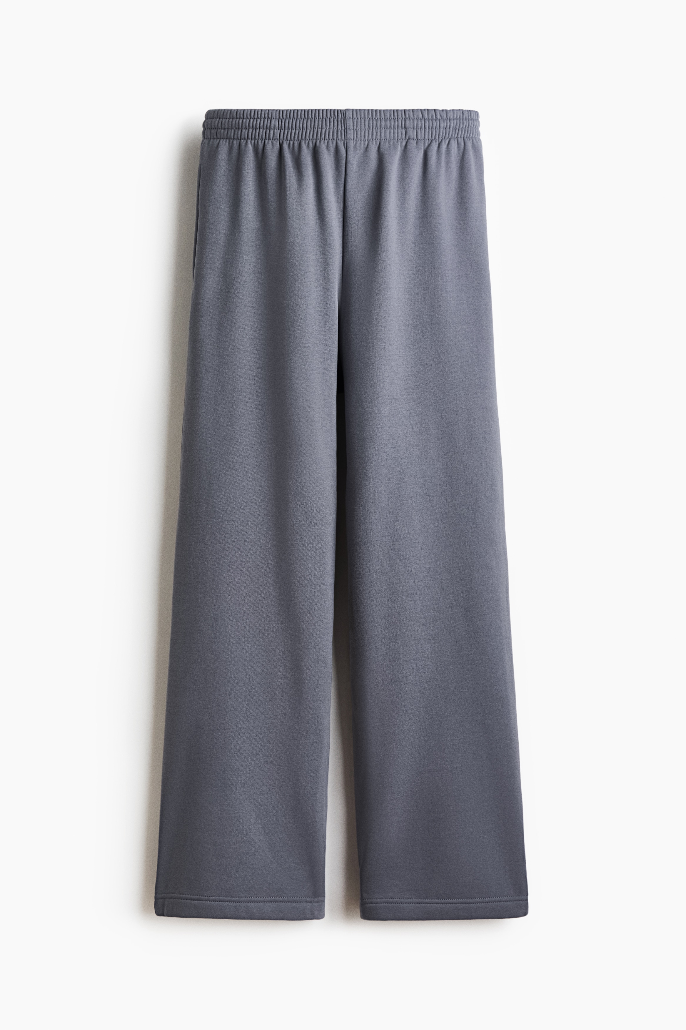 Track pants in a muted blue-grey sweat fabric are worn by the model, featuring a relaxed straight-leg fit and a drawstring waist. The pants are styled with a white long-sleeve top, a matching sweatshirt draped over the model's shoulder, and black flat sandals.