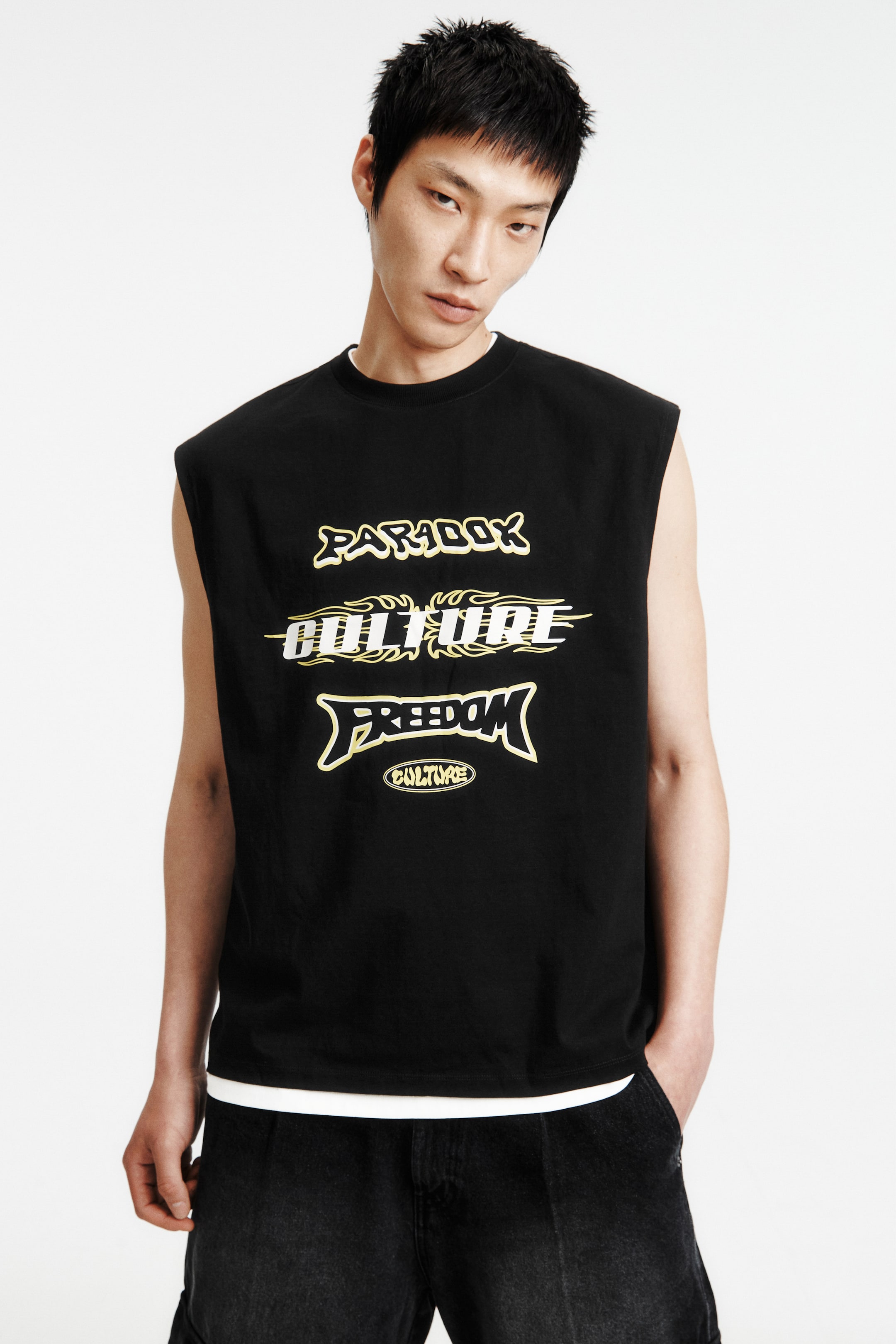 View larger image: A black sleeveless top featuring a crew neck and a striking graphic print on the front. The print showcases the words "PARADOX," "CULTURE," and "FREEDOM" in stylized yellow and white text, with a smaller "CULTURE" graphic beneath. It is layered over a white t-shirt, with the white hem visible.