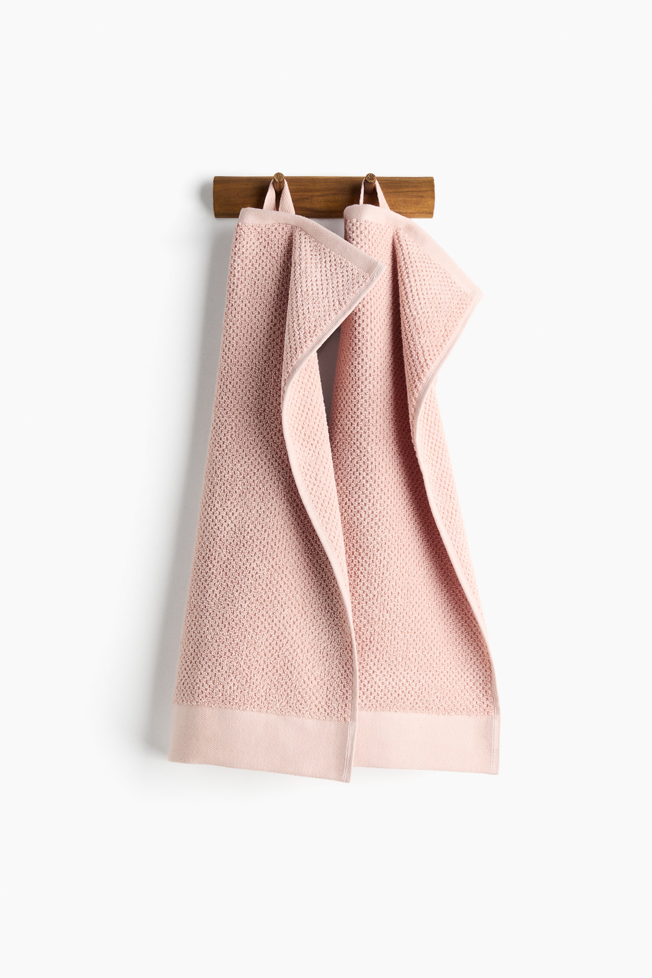 View larger image: Two light pink guest towels are hanging by loops located on one short side, each displaying a textured, small-weave pattern with smooth, flat borders along its edges and draped.