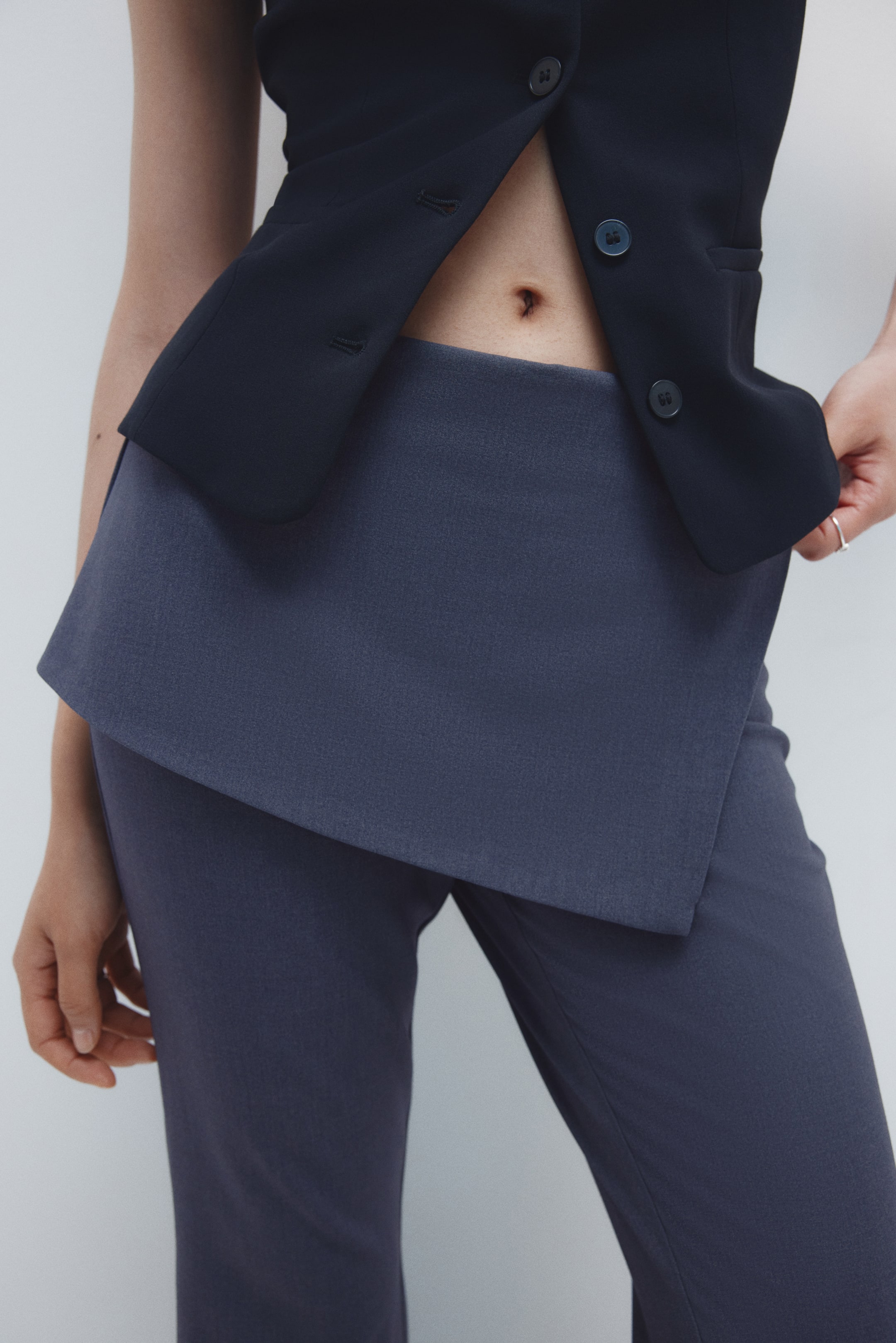 View larger image: A pair of dark blue trousers, viewed from the front-side, is featuring an asymmetrical, overlapping fabric panel layered over the waist, extending over the upper thighs. A dark navy button-front vest is being worn unbuttoned, revealing the midriff and partially covering the layered waist detail of the trousers.
