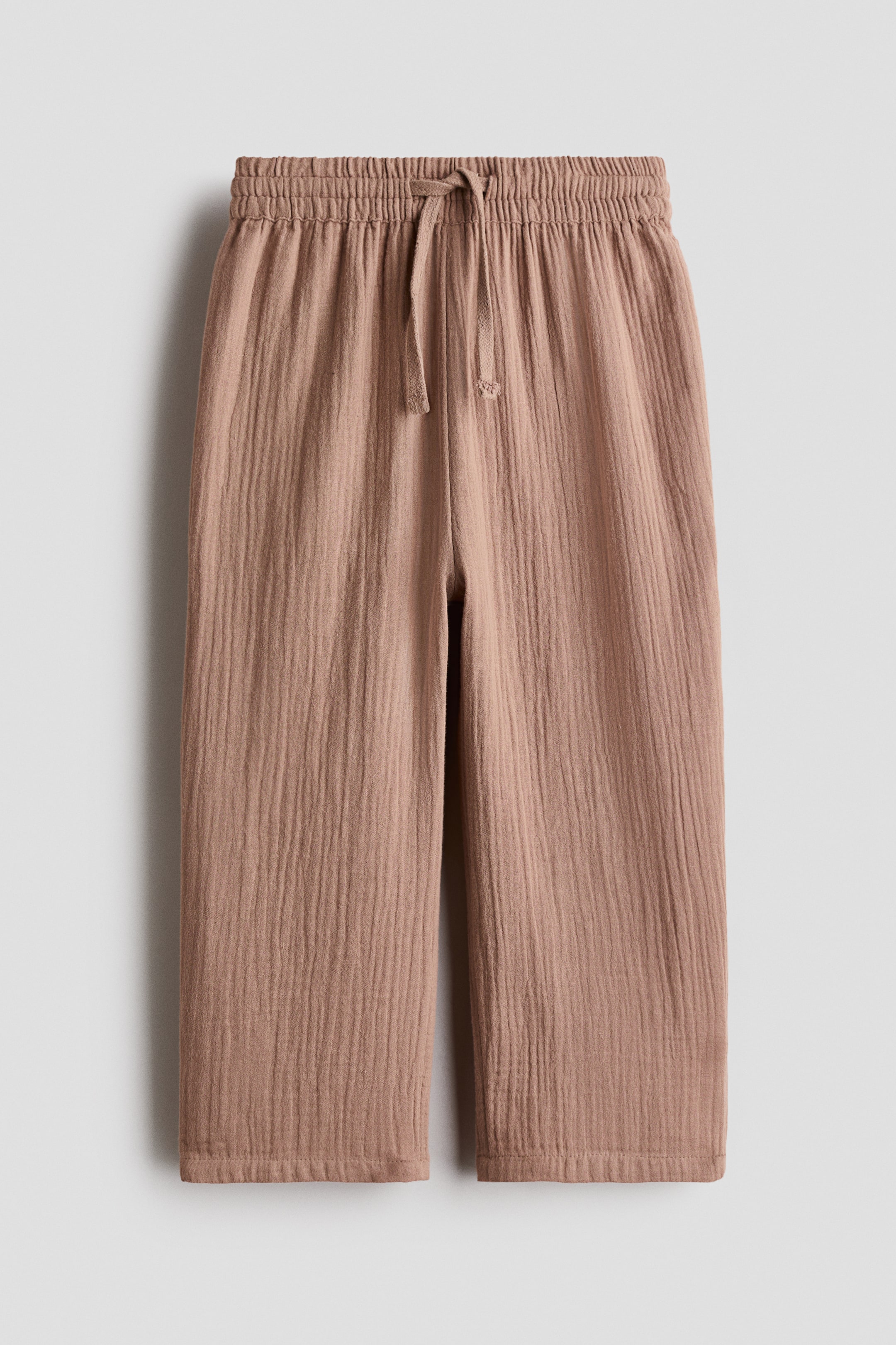 View larger image: Light brown muslin joggers are showcased from the front, featuring an elasticated waist with a tied drawstring and straight, relaxed legs with a visible crinkled texture.