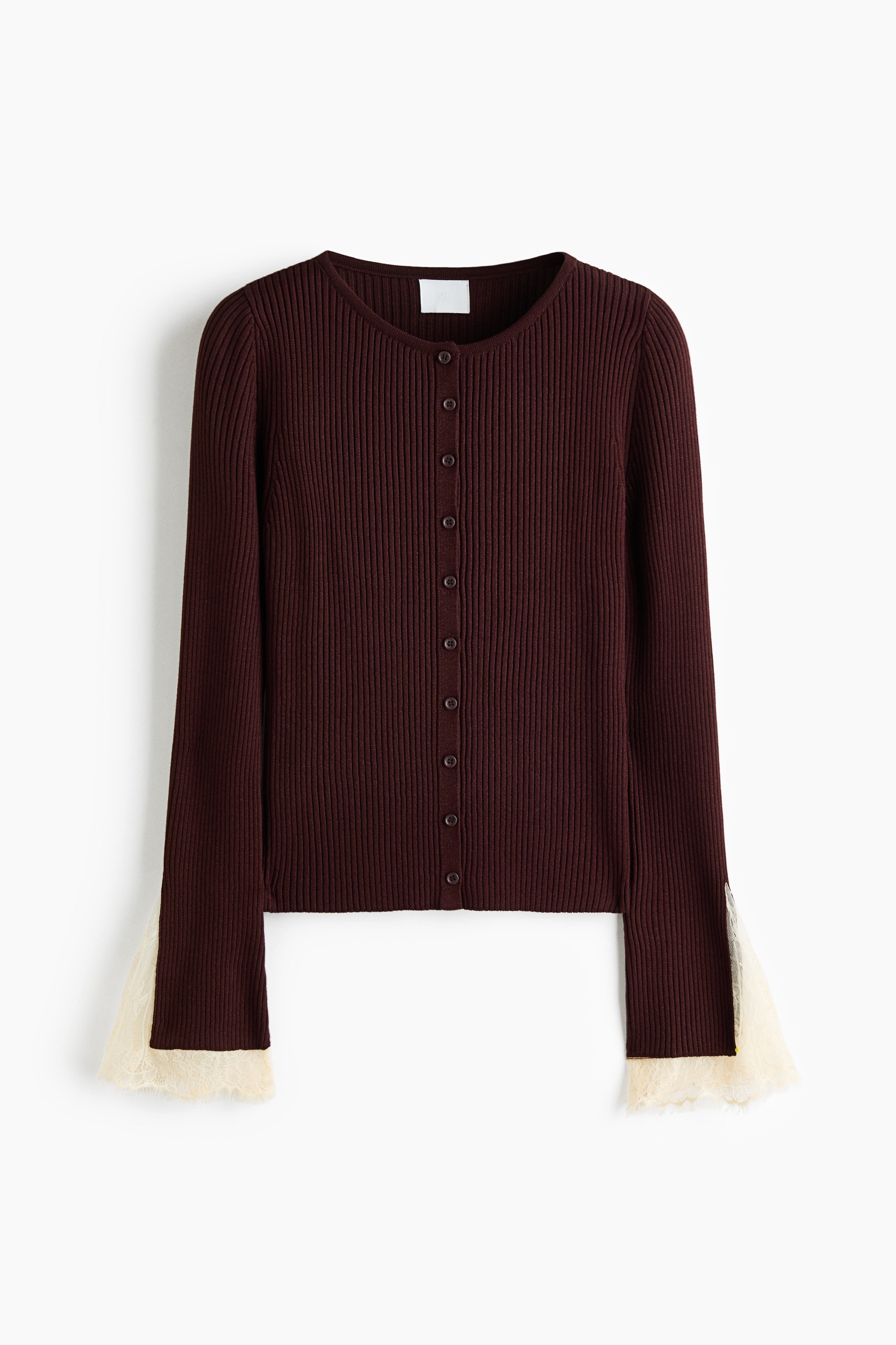A dark brown ribbed knit cardigan with a button-down front is being worn by the model, who has her arms crossed to showcase the flared cuffs trimmed with cream-coloured lace. The cardigan is styled with off-white trousers and a dark belt.