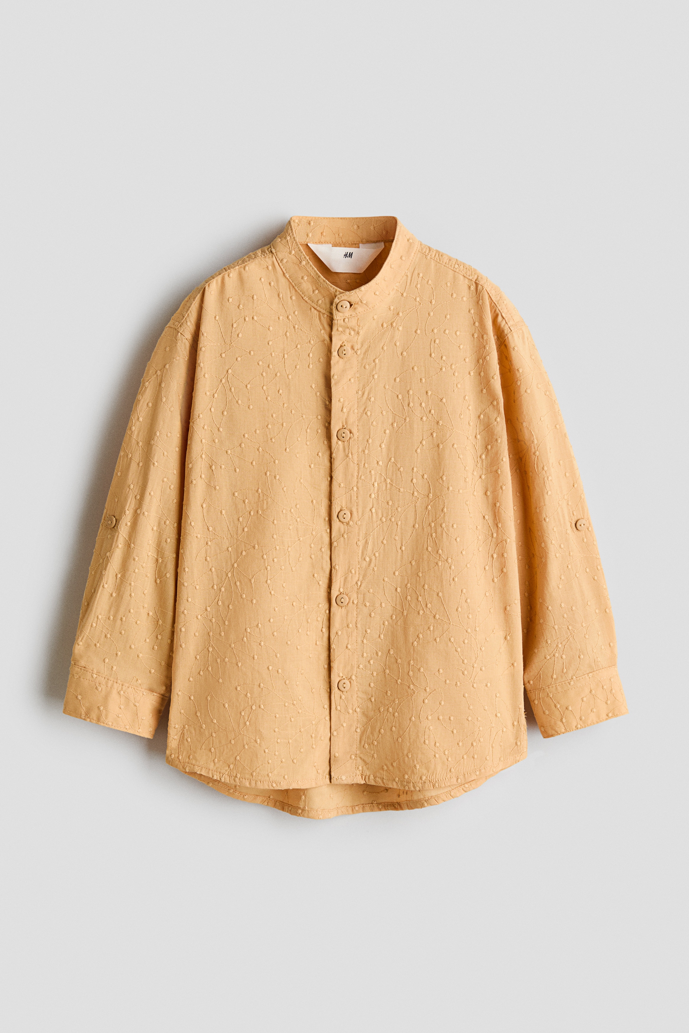 A front view shows a light apricot-coloured grandad shirt in a soft, patterned cotton weave, featuring a band collar and a button-down front. The long sleeves with button cuffs are being rolled up, and the shirt is finished with a gently rounded hem.