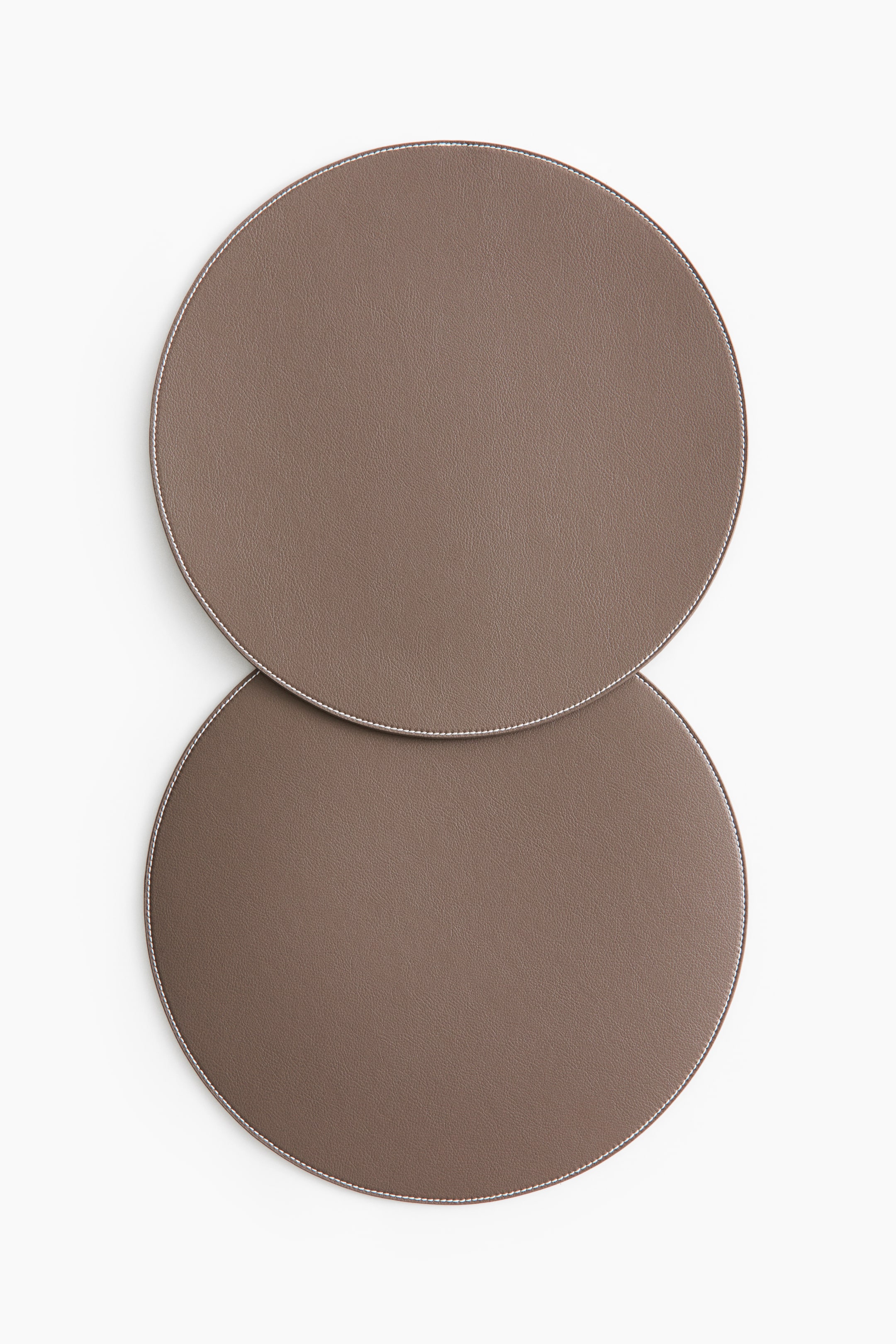 View larger image: Two round placemats are showcased, crafted from brown coated fabric. Each placemat highlights contrasting white stitching along its edge, with one placemat partially overlapping the other.
