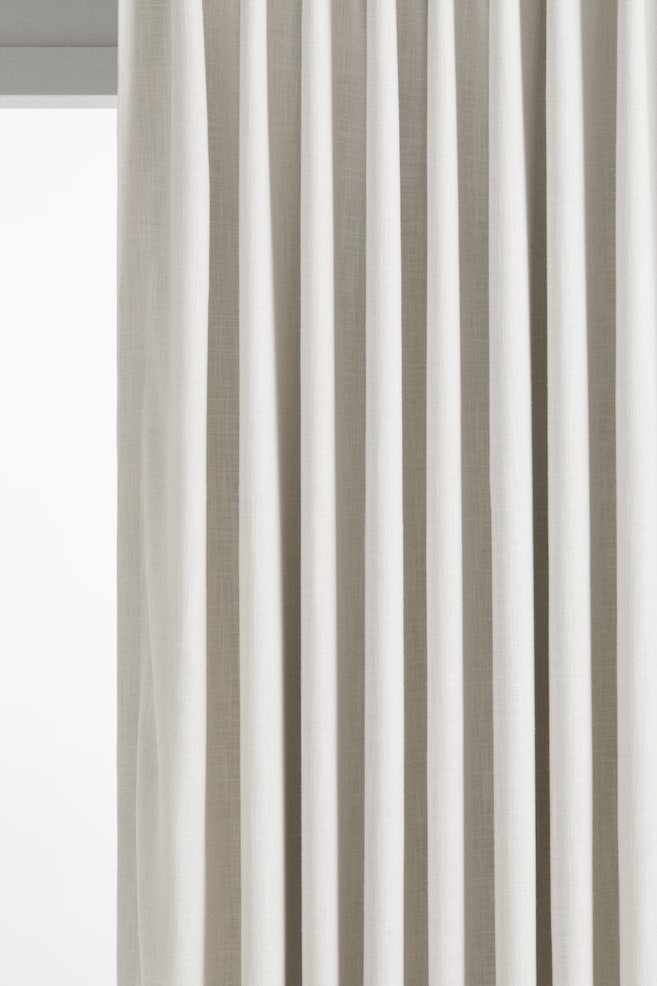 Vista frontal aproximada de um cortinado bege-claro suspenso em elegantes pregas verticais. O tecido apresenta uma trama texturada de linho e viscose com um brilho subtil, exibindo um caimento fluido. - 1