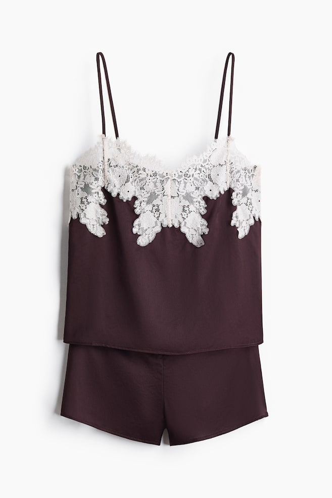 A dark plum satin pyjama set is hanging in a front view, showcasing a camisole with thin spaghetti straps and intricate white floral lace trim along the neckline, paired with matching satin shorts. - 1