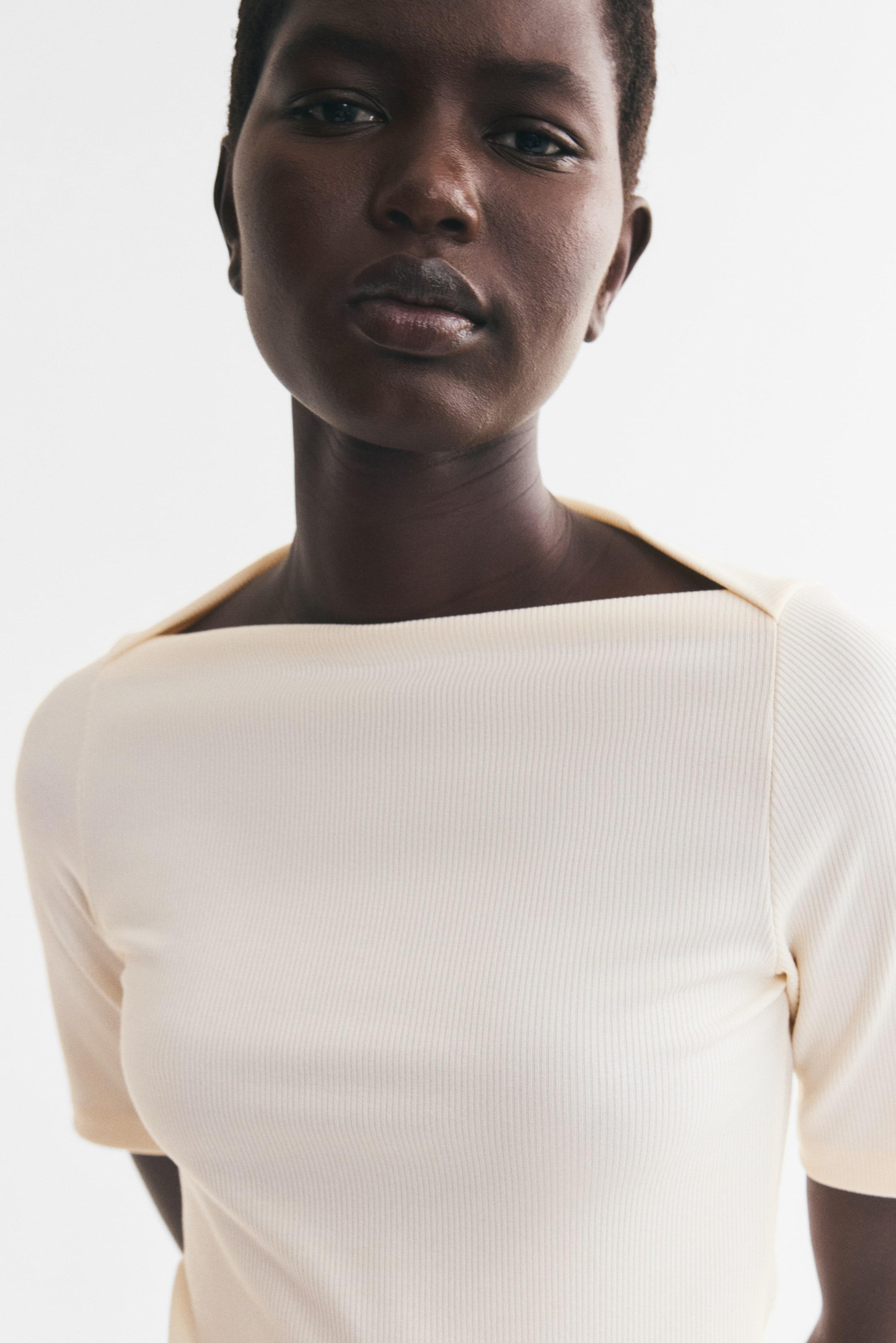 View larger image: A cream-colored ribbed-knit short-sleeve top is being worn by the model, showcasing its fitted silhouette. The top features a wide boat neckline with subtle overlap details at each shoulder.