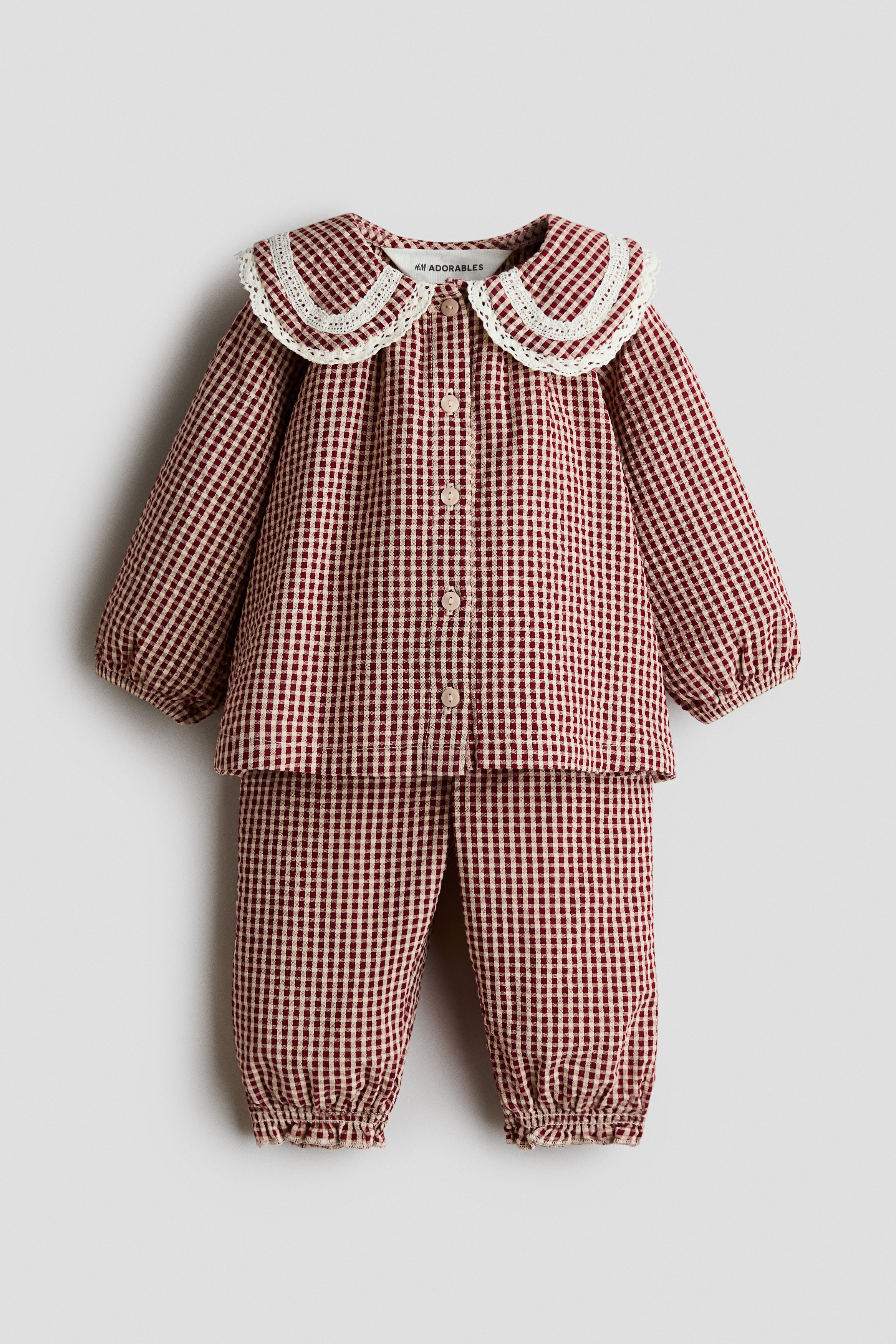 KÄ±rmÄ±zÄ± ve beyaz gingham desenli, uyumlu iki parÃ§alÄ± pamuklu bir bluz ve pantolon takÄ±mÄ± Ã¶nden gÃ¶sterilmektedir; geniÅ, yuvarlak, dantelli yakaya sahip, uzun kollu, Ã¶n kÄ±smÄ± dÃ¼Ämeli ve manÅetleri lastikli bir bluz iÃ§ermektedir. TakÄ±mÄ± tamamlayan bol kesim pantolonlar ise bileklerde lastikli, fÄ±rfÄ±r detaylÄ± paÃ§alarla sunulmaktadÄ±r.
