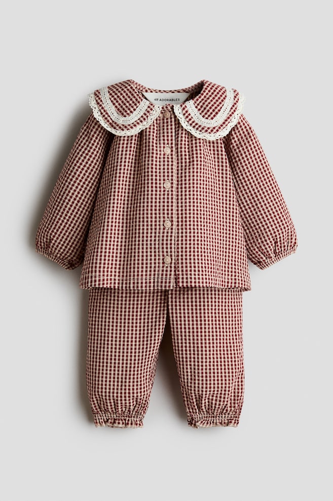 A red and white gingham blouse and pants set is displayed from a front angle. The blouse is featuring a wide, rounded collar with white lace trim, a button-down front, and elasticated cuffs, while the matching pants are showing elasticated, frill-trimmed hems. - 1