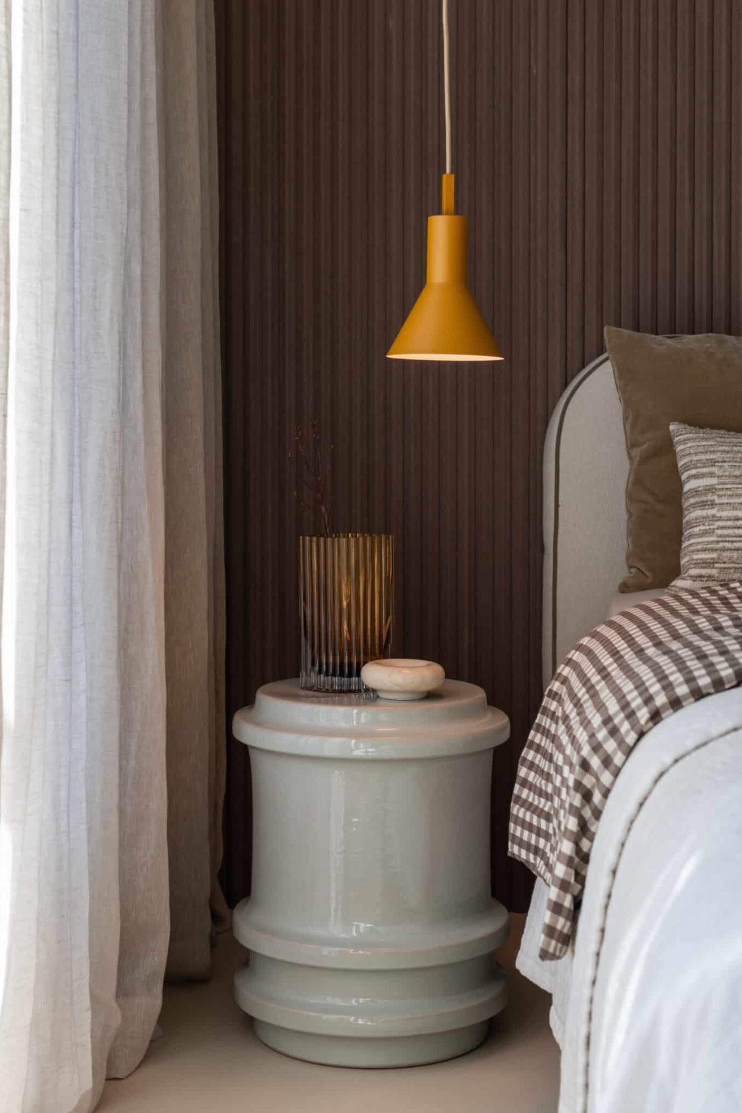 De hanglamp hangt verticaal en toont een matte okerbruine afwerking met een cilindrisch bovenstuk dat overgaat in een brede kegelvormige kap. Een vierkant blok verbindt de kap met een gevlochten stoffen snoer, waarbij de warme, lichtgekleurde binnenvoering zichtbaar is.