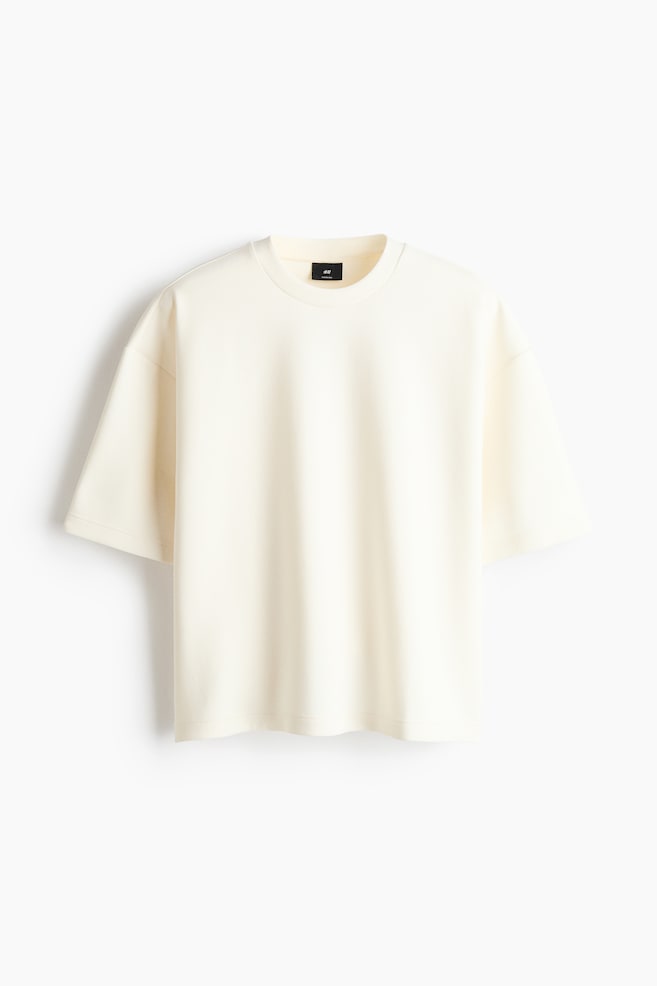 An off-white T-shirt is facing front, featuring a relaxed fit with dropped shoulders, short sleeves, a ribbed crew neck, and a straight hem. - 1