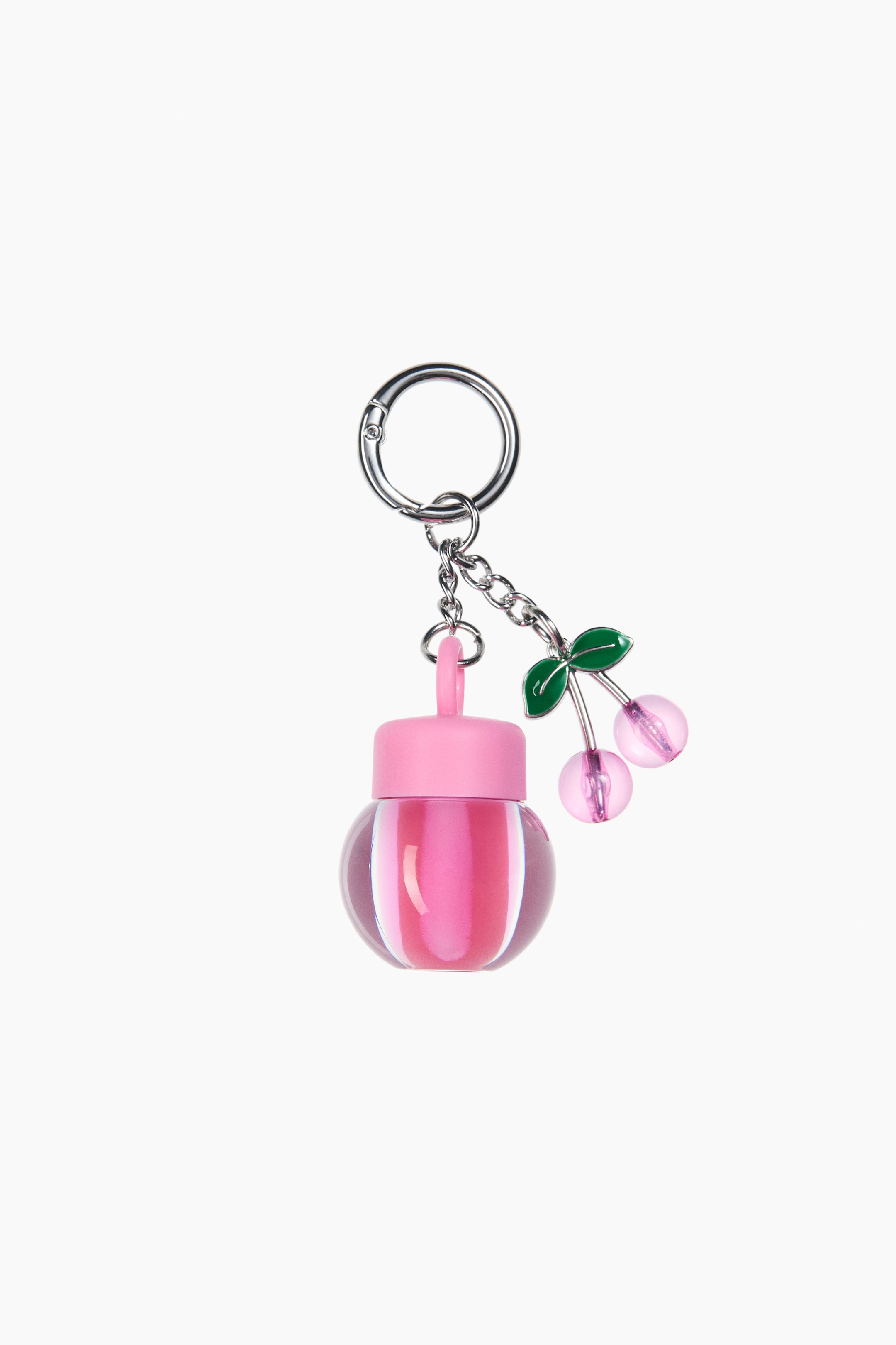 View larger image: The bag charm showcases a clear spherical container filled with pink lip oil, capped with a pink lid. It hangs from a silver-toned carabiner clasp via a chain, which also supports a pink cherry-shaped pendant adorned with green leaves.