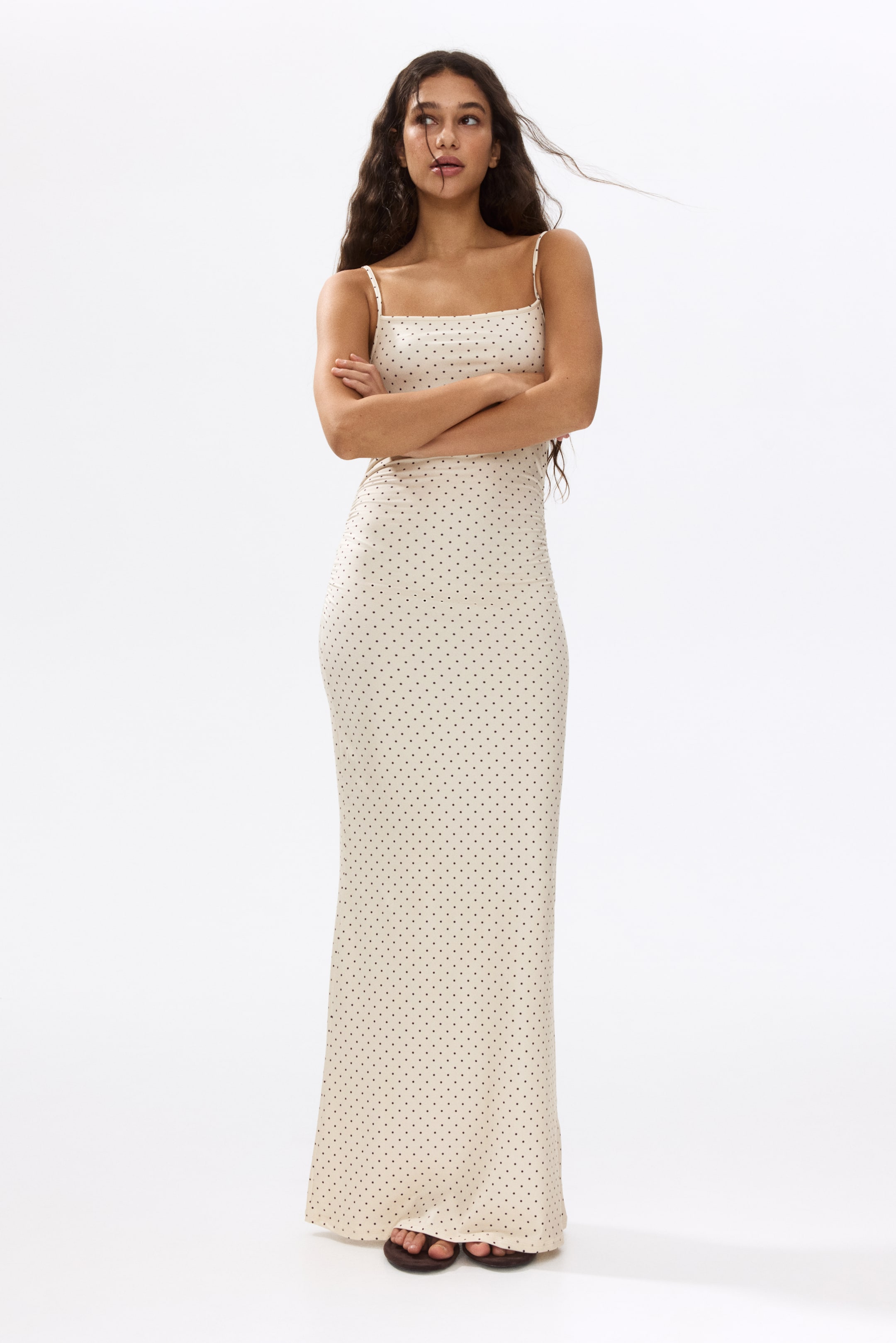 拡大画像を表示: クリーム色のマキシドレスは、薄いスパゲッティストラップとスクエアネックラインを特徴とするフルフロントビューで紹介されています。体にぴったりとフィットし、全体に小さな黒いポルカドットのパターンが施されています。