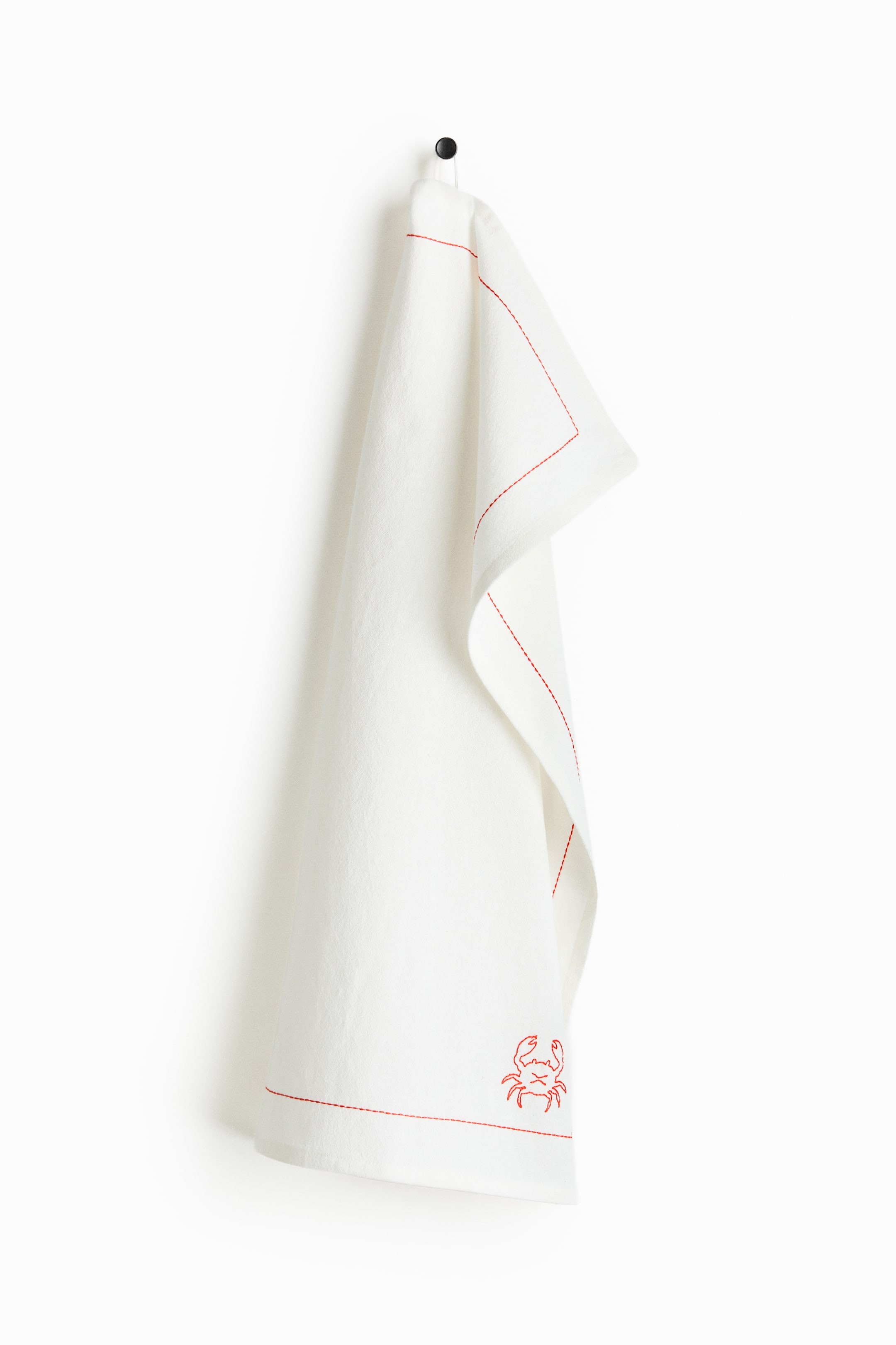 Visualizza immagine più grande: "Un canovaccio color crema, tessuto con una miscela di cotone e lino, è appeso per un anello a un gancio nero, con contorni ricamati in rosso lungo i bordi e un motivo di granchio nell'angolo in basso a destra."