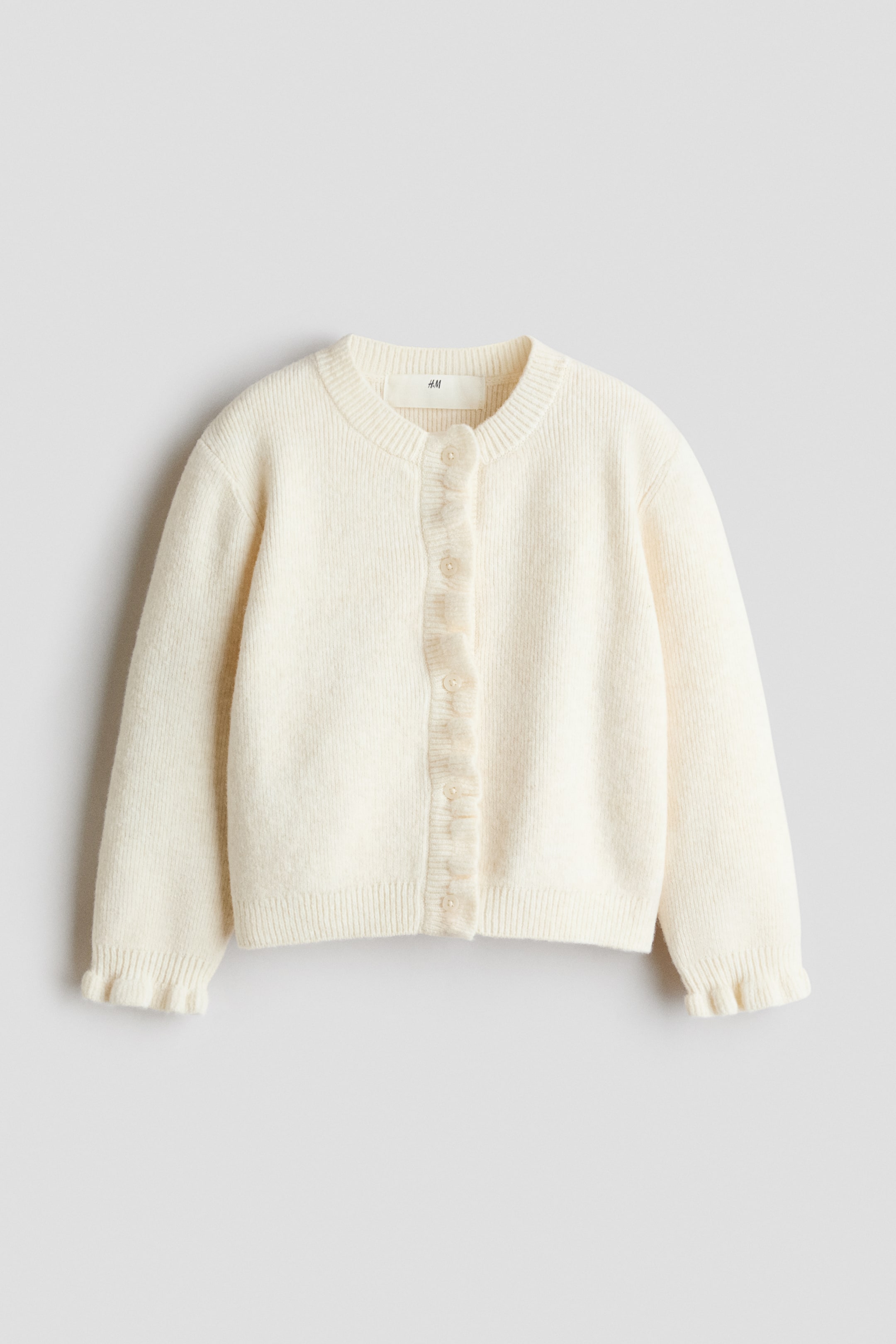 View larger image: A cream cardigan is shown from the front, featuring a round ribbed neckline and a button-front fastening with small, light-coloured buttons. It has a ruffled placket down the front and ruffled cuffs on its long sleeves.