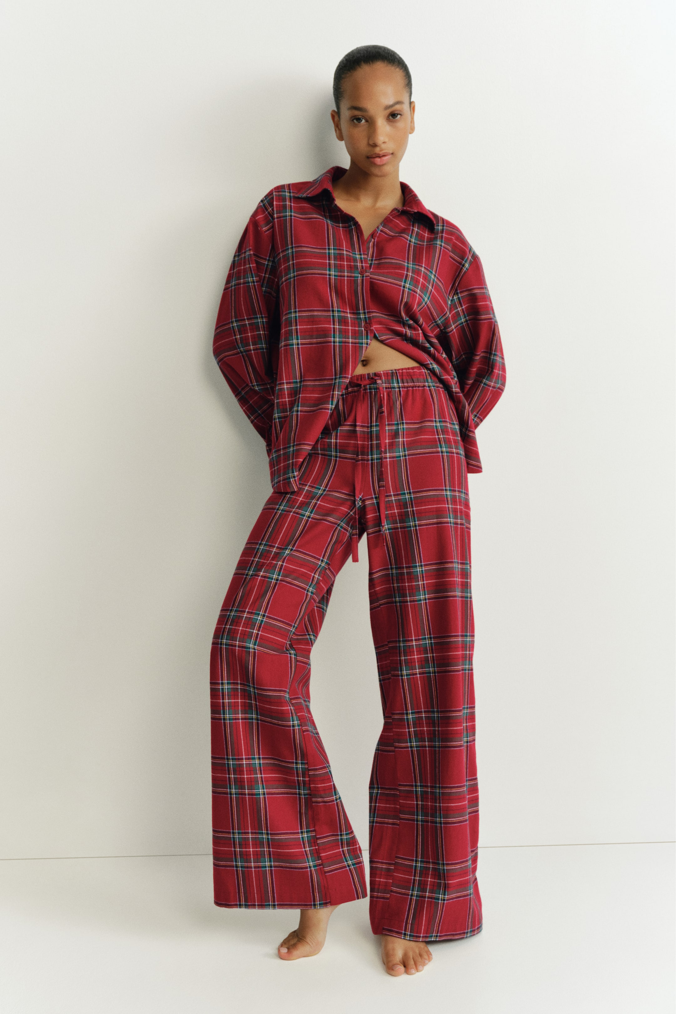 View larger image: A red plaid flannel pyjama set is worn by the model, featuring a long-sleeved, collared button-down shirt and matching wide-leg pants. The shirt is left partially unbuttoned at the lower hem, revealing the elasticated drawstring waistband of the pants.