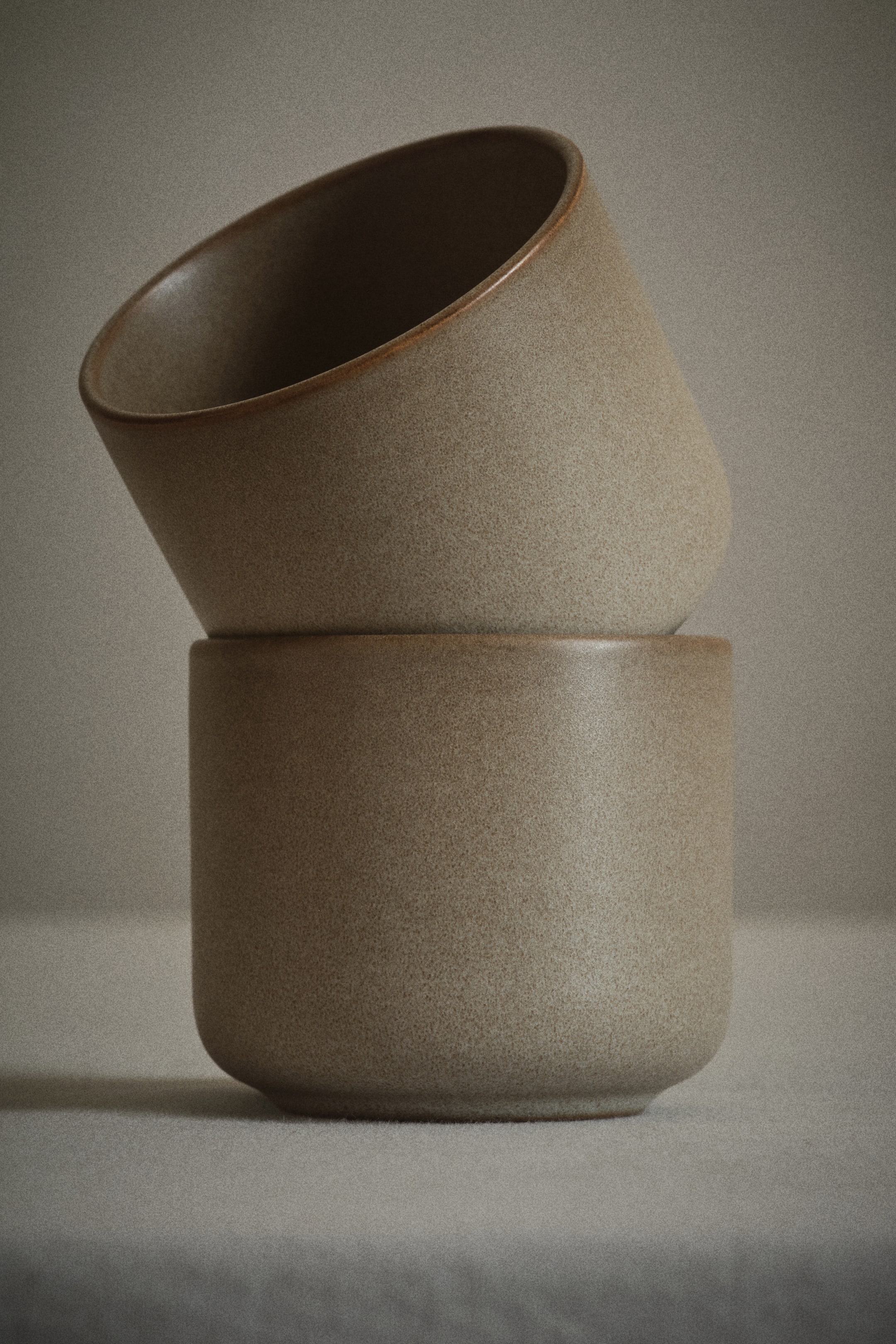 View larger image: Two speckled beige ceramic cups are stacked one on top of the other. The upper cup, with a slightly flared rim and a deeper brown interior, is tilted and resting on its side rim on the upright lower cup, which shows a rounded base.