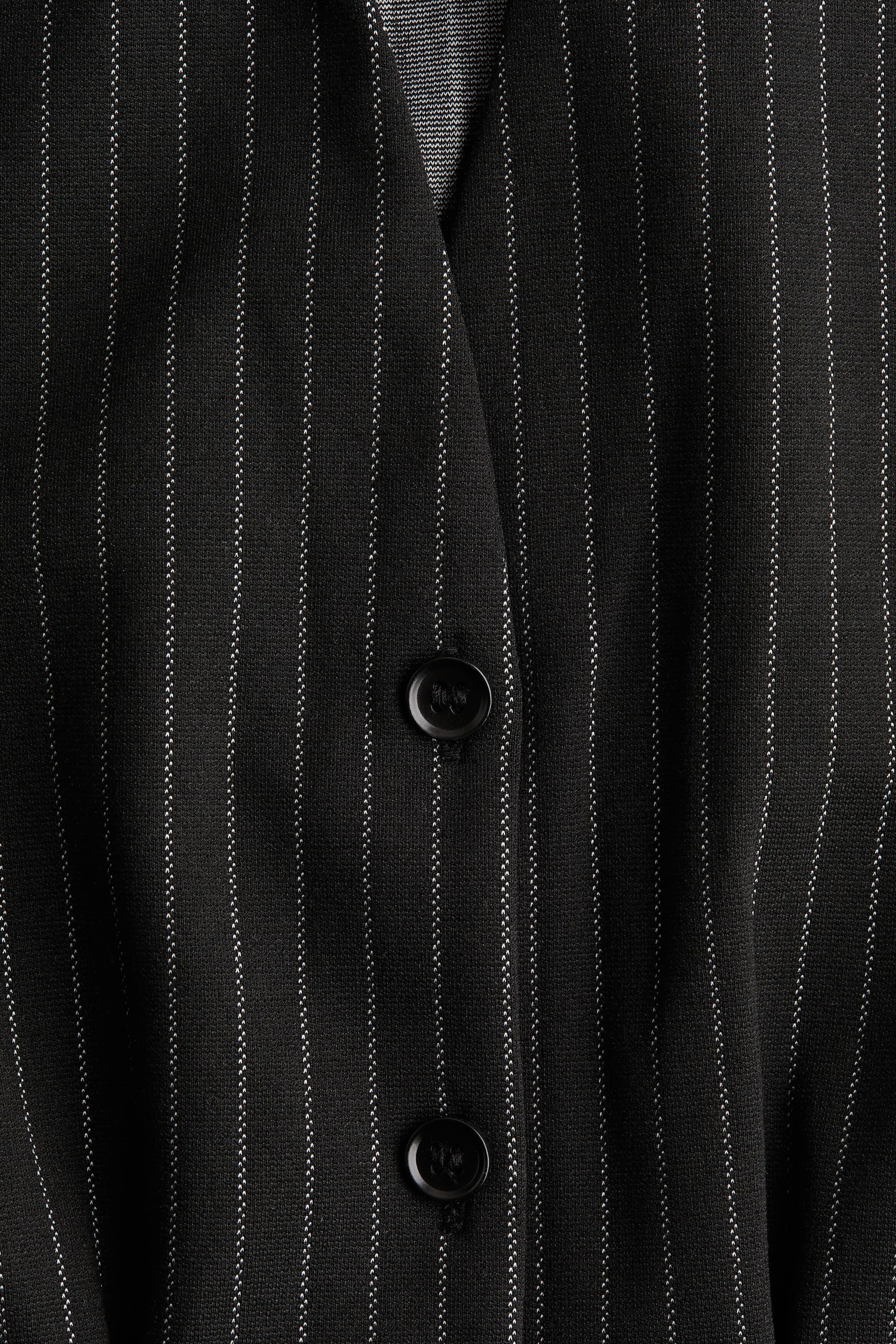 View larger image: The front of a black pinstripe garment is being shown, featuring thin white vertical pinstripes. Two round black buttons are visible on the front placket, with a structured V-shaped opening at the top.