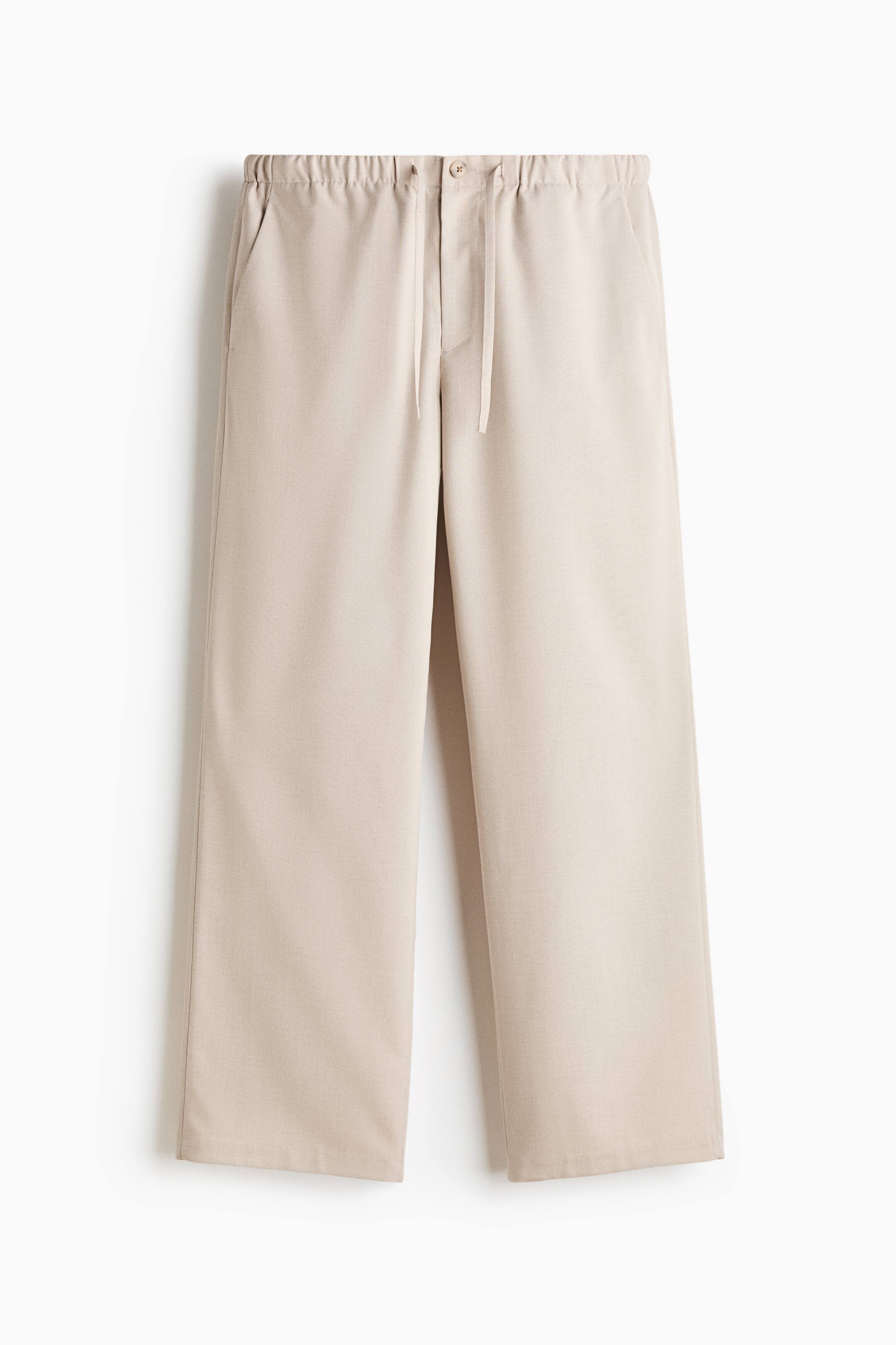 View larger image: A pair of light beige trousers is shown from the front, featuring an elasticised waistband with a matching drawstring and a single button closure. The trousers fall into a relaxed, wide-leg silhouette with side seam pockets and straight hems.