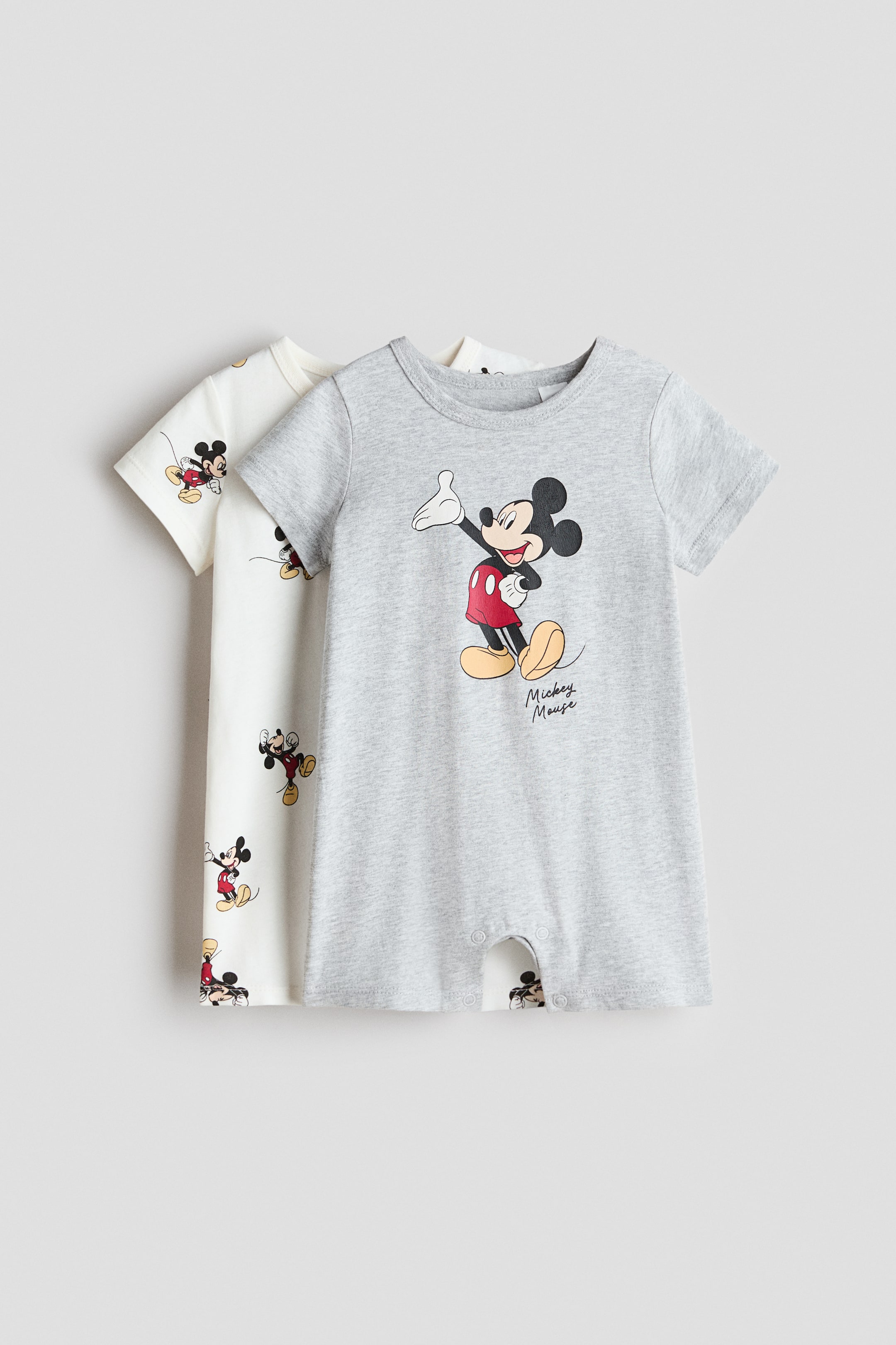 View larger image: Two short-sleeved sleepsuits are showcased at the front, crafted from soft, printed cotton jersey. The front grey sleepsuit features a large Mickey Mouse graphic, while the partially visible off-white sleepsuit behind it boasts an all-over Mickey Mouse print. Both sleepsuits are designed with a round neckline, short sleeves, short legs, and press-stud fastenings at the crotch.