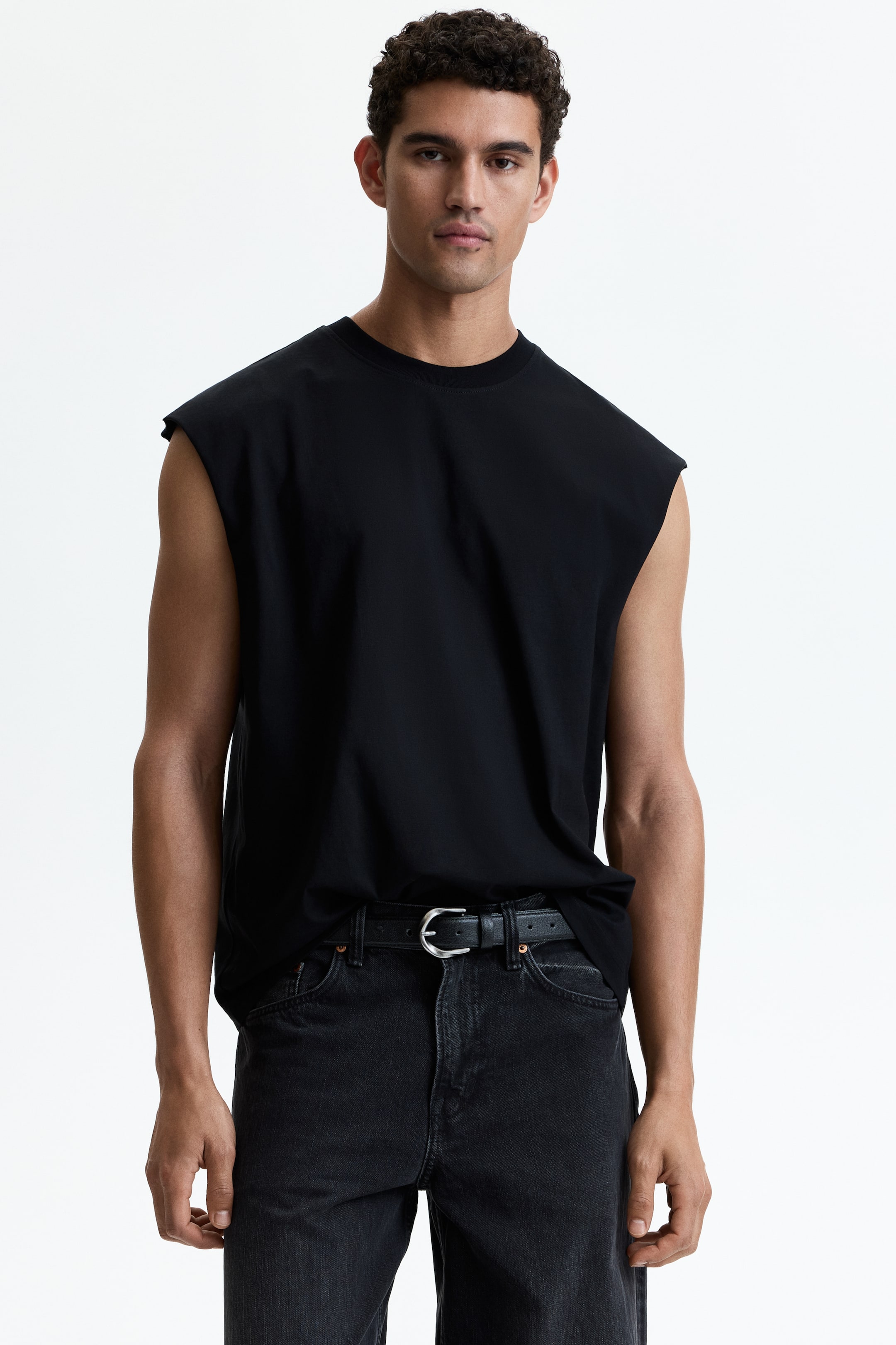 Visa större bild: En svart ärmlös t-shirt visas framifrån, med en rund hals och förlängda axellinjer, vilket skapar en bred, muskel-t-shirt silhuett. Den har en avslappnad passform, med fållen delvis instoppad på höger sida medan vänster sida hänger löst.