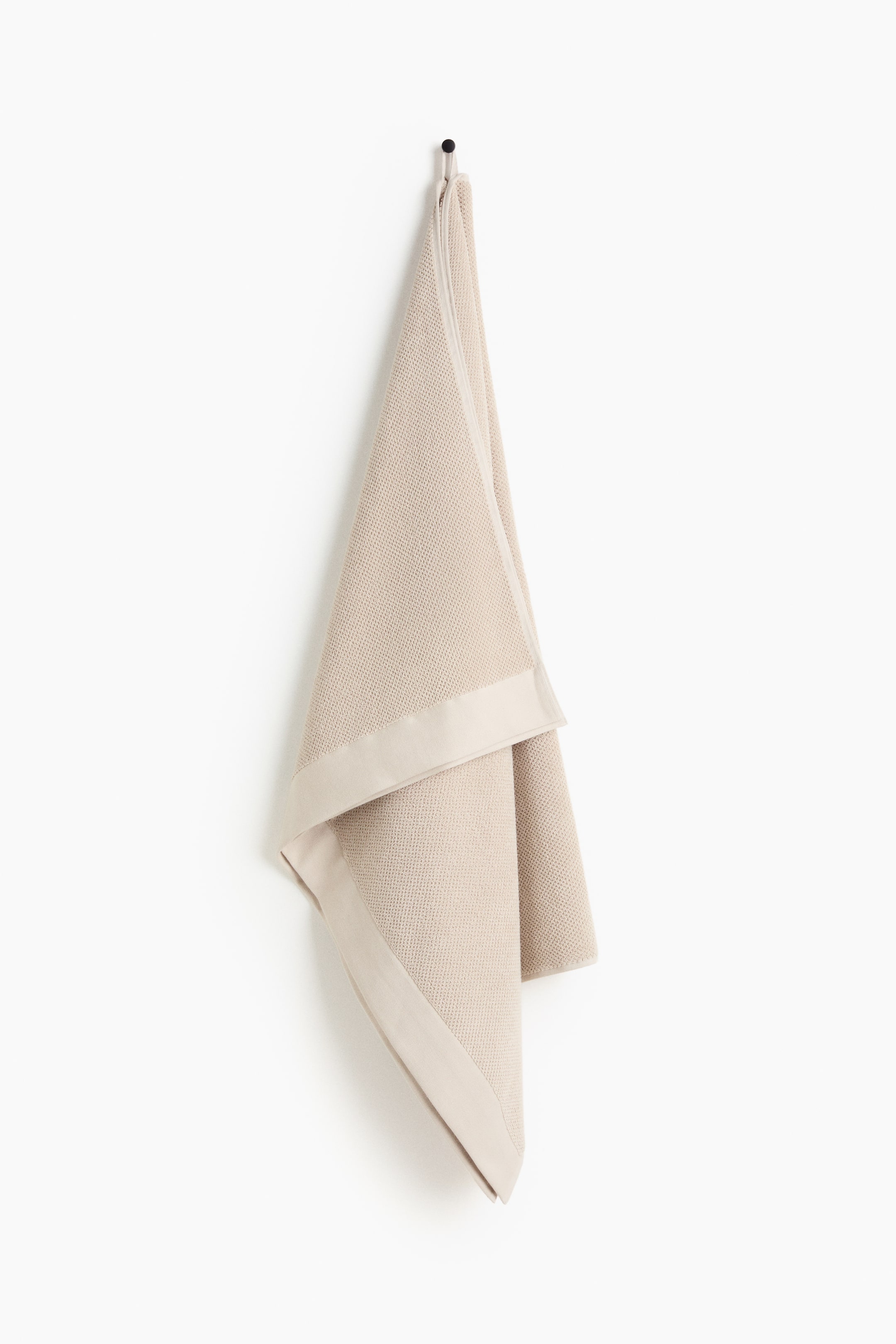 View larger image: A light beige classic bath sheet is hanging folded from a black hook, showcasing a subtle woven texture and a smooth, slightly contrasting border along its edges.