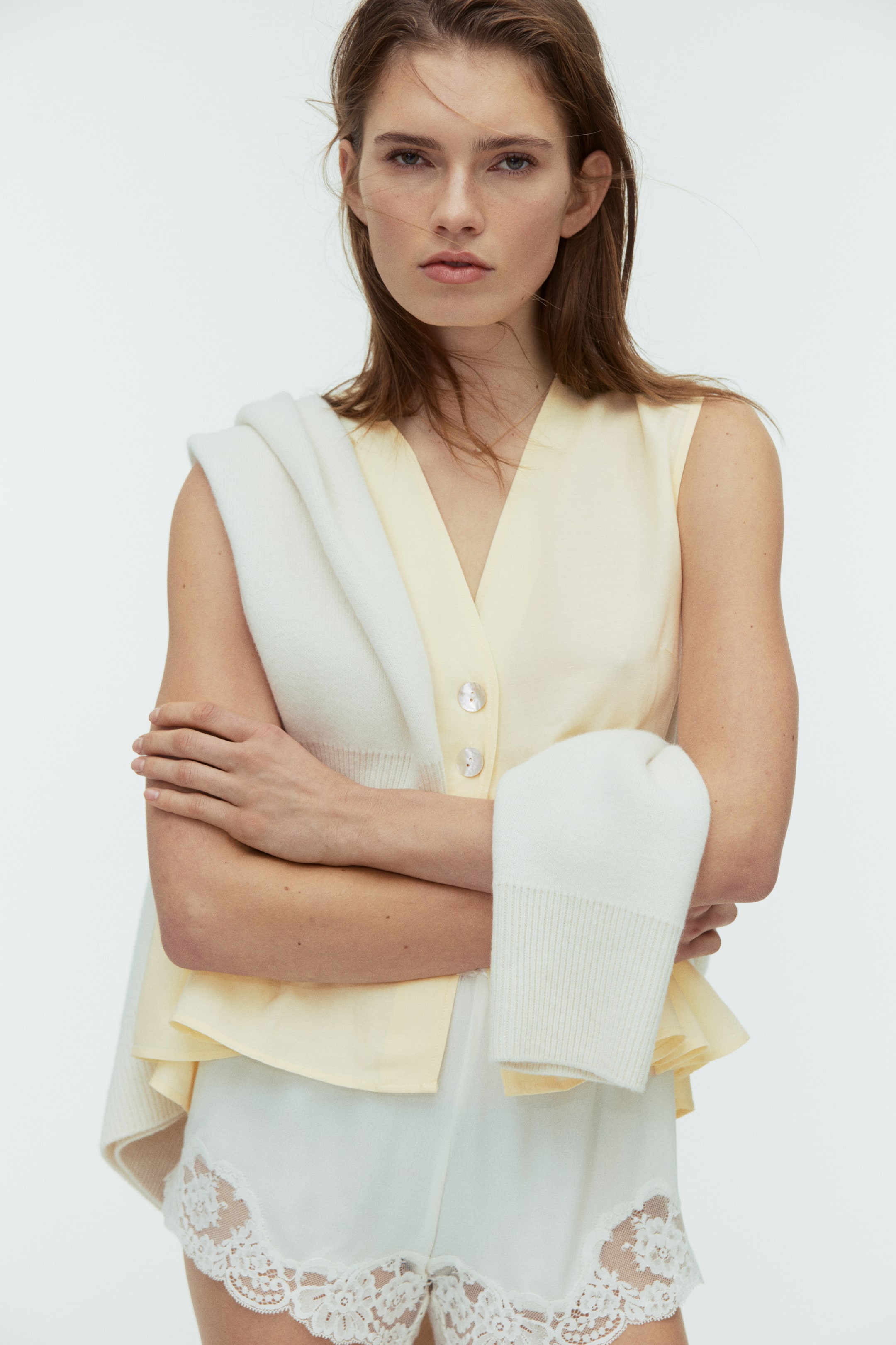 View larger image: A pale yellow sleeveless top features a V-neckline and a button-front closure with two visible iridescent buttons. The front view shows a flared peplum hem, and the top is partially covered by a white cardigan draped over the shoulders.