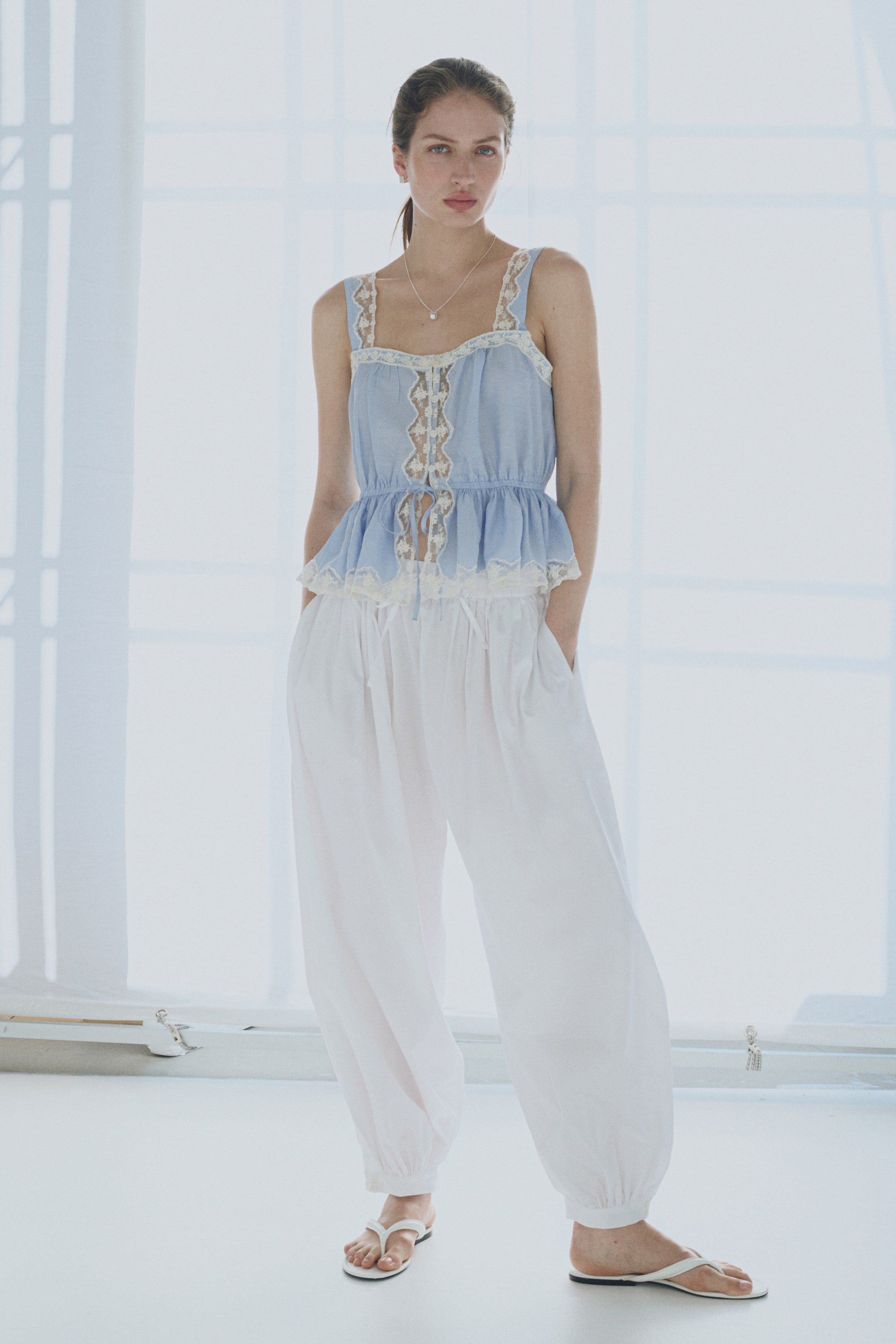 View larger image: The model is wearing a light blue woven fabric top adorned with delicate lace trims along the edges. It features a square neckline and wide shoulder straps. The top includes button and loop fasteners down the front, an elasticated seam at the waist with narrow ties, and a flared peplum.