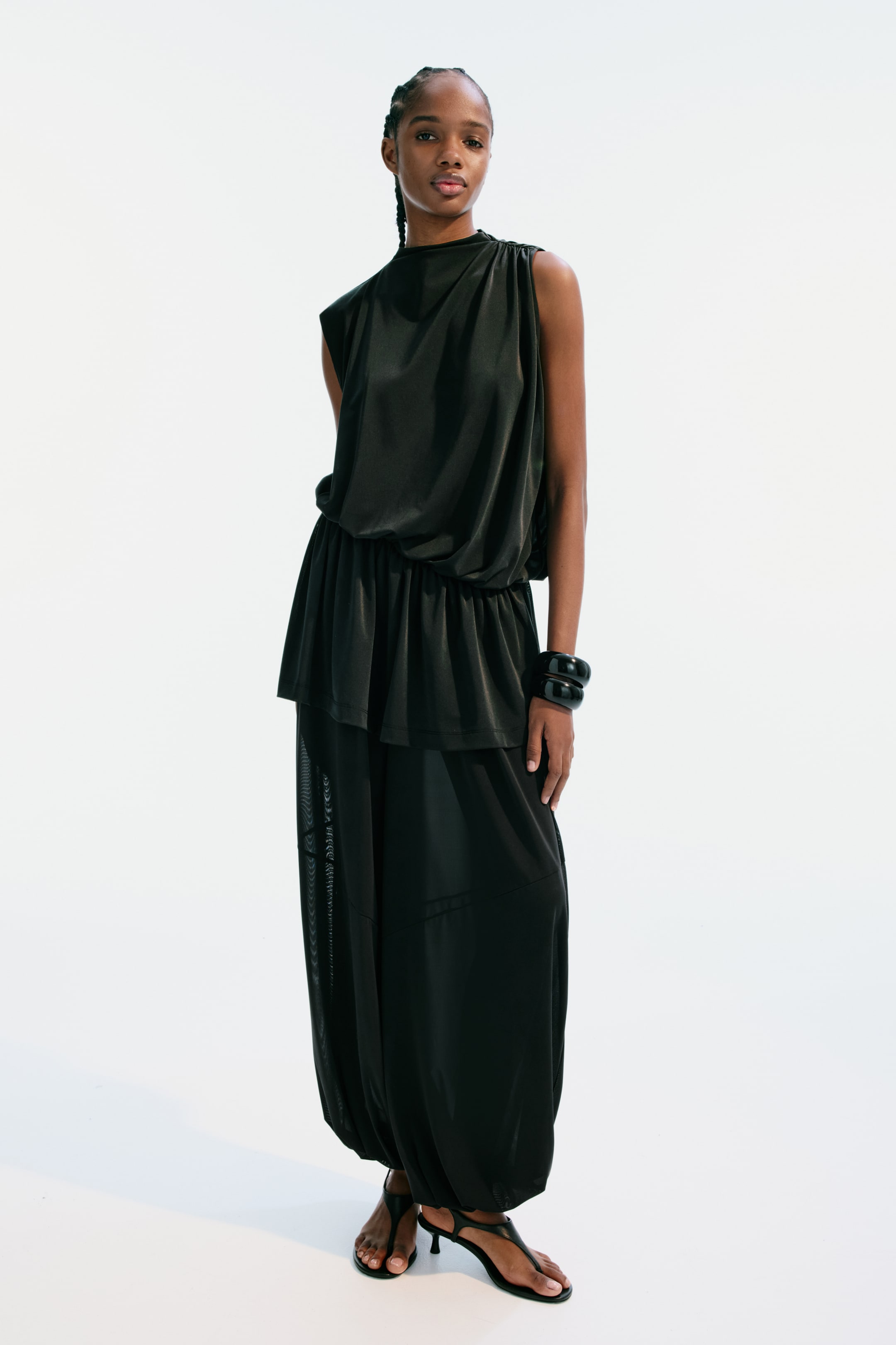 View larger image: The model is wearing a black sleeveless top in stretch jersey, featuring a stand collar, gathers on the shoulders, and an asymmetric hem. Its loose-fit bodice with an elasticated seam at the waist creates a slouchy, draped effect.