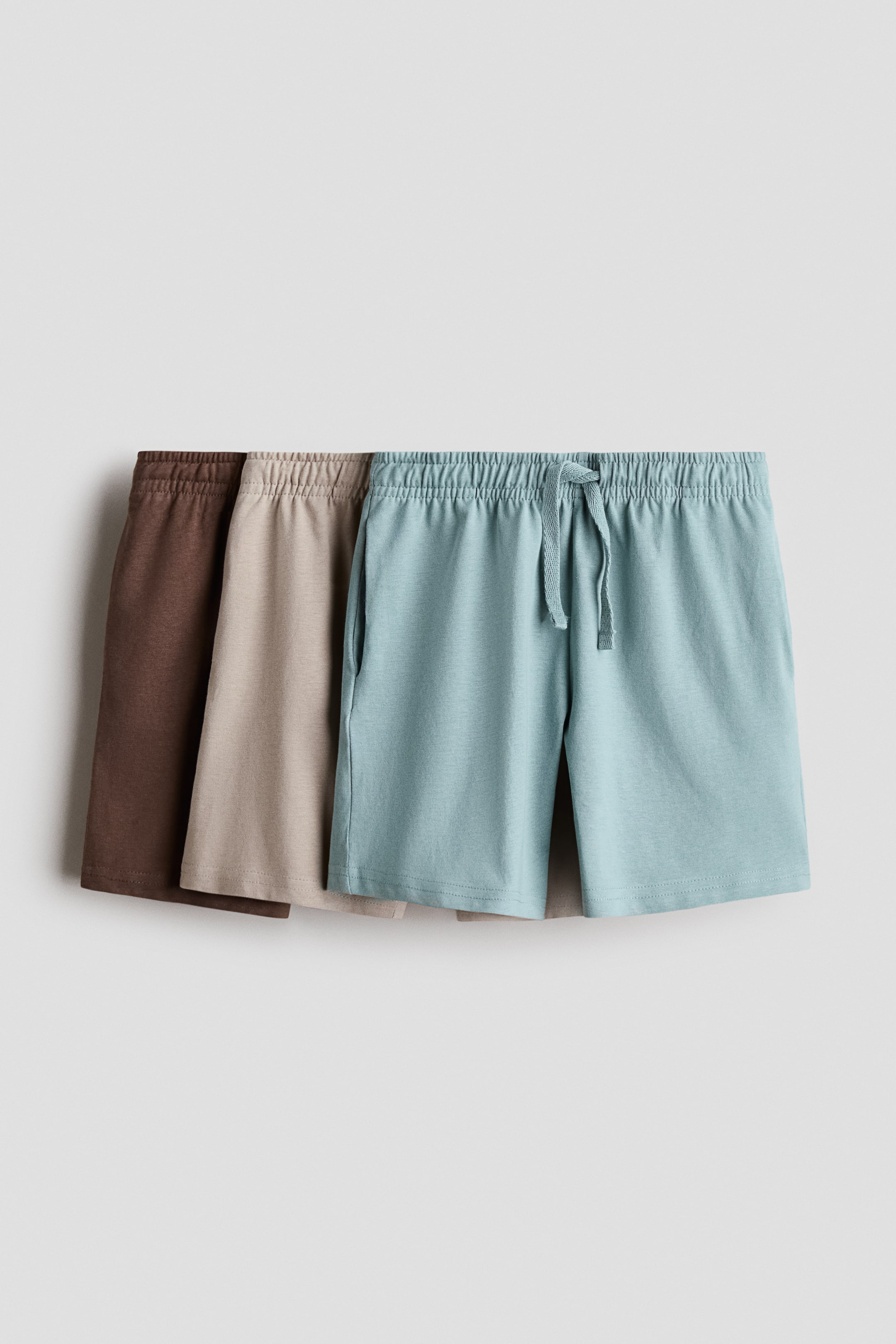 View larger image: Three pairs of soft cotton jersey shorts are displayed, each featuring an elasticated, drawstring waist. The front light blue pair showcases a tied drawstring and side pockets, with a beige and a brown pair partially visible behind it.