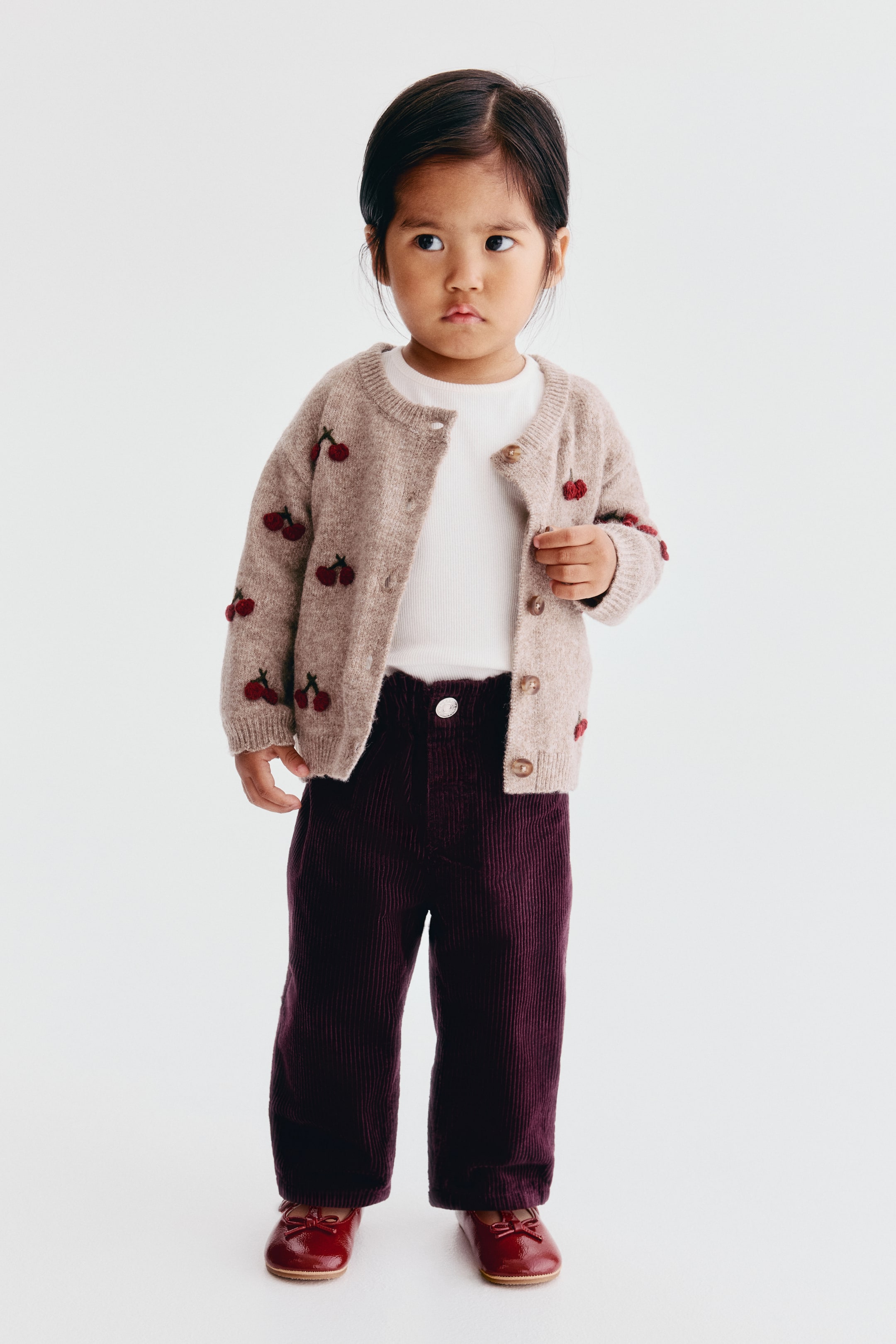 View larger image: The dark purple corduroy trousers feature a straight-leg design with an adjustable, elasticated paperbag waist and a mock fly with a single button. A white T-shirt is tucked into the trousers to showcase the gathered waistline and decorative front pockets from a front-facing angle.