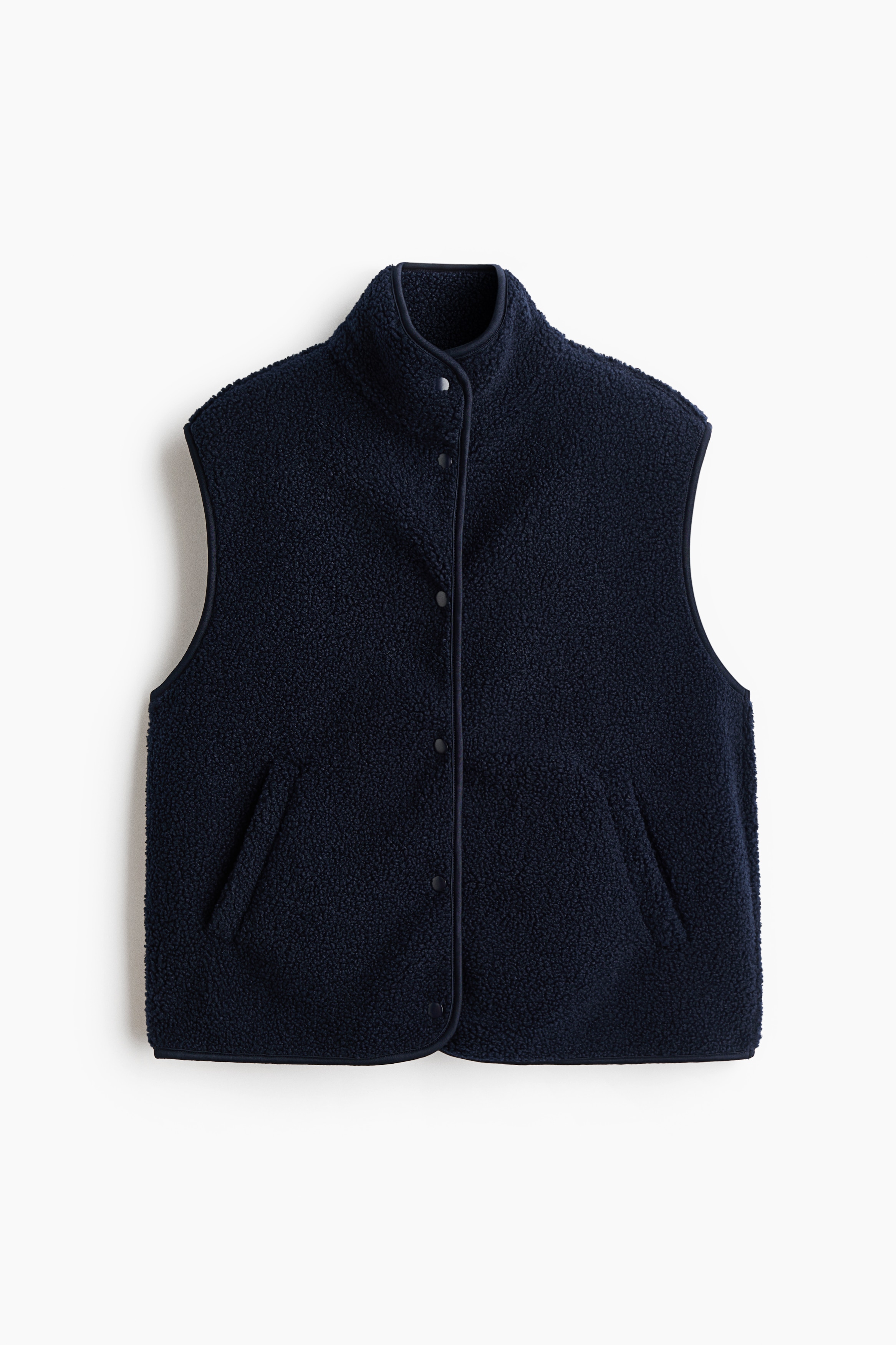 A navy blue teddy fleece vest is being worn by the model over a matching long-sleeve hooded sweatshirt and skirt. The vest features a high funnel collar, jersey-bound edges, and diagonal front pockets in which the model is resting their hands.