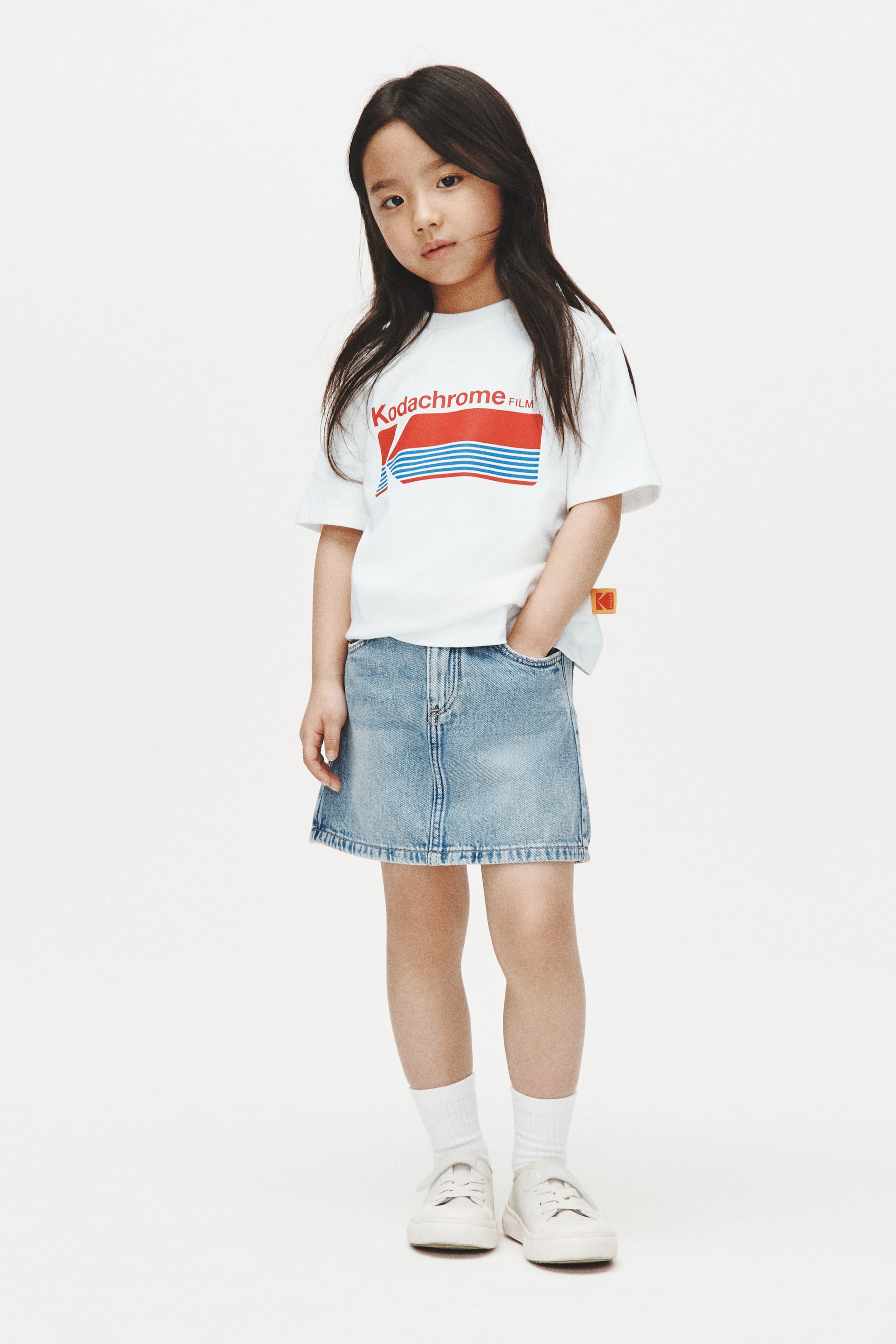 拡大画像を表示: 白の半袖Tシャツに、赤と青の「Kodachrome FILM」グラフィックが前面に施されており、ライトブルーのデニムミニスカートに部分的にインして着用されています。スカートには前ポケットとステッチのディテールが見えます。