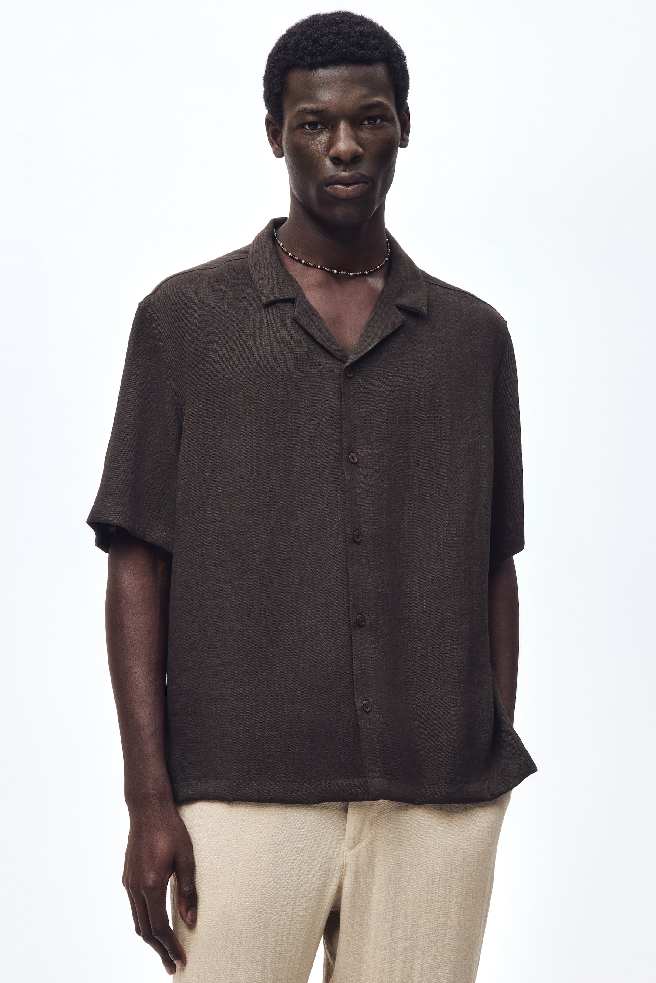 View larger image: The model is wearing a dark brown short-sleeved shirt crafted from a textured viscose-blend weave. It features a resort collar, French front, and a straight-cut hem. The shirt offers a relaxed fit and is paired with light-colored trousers and a beaded necklace.