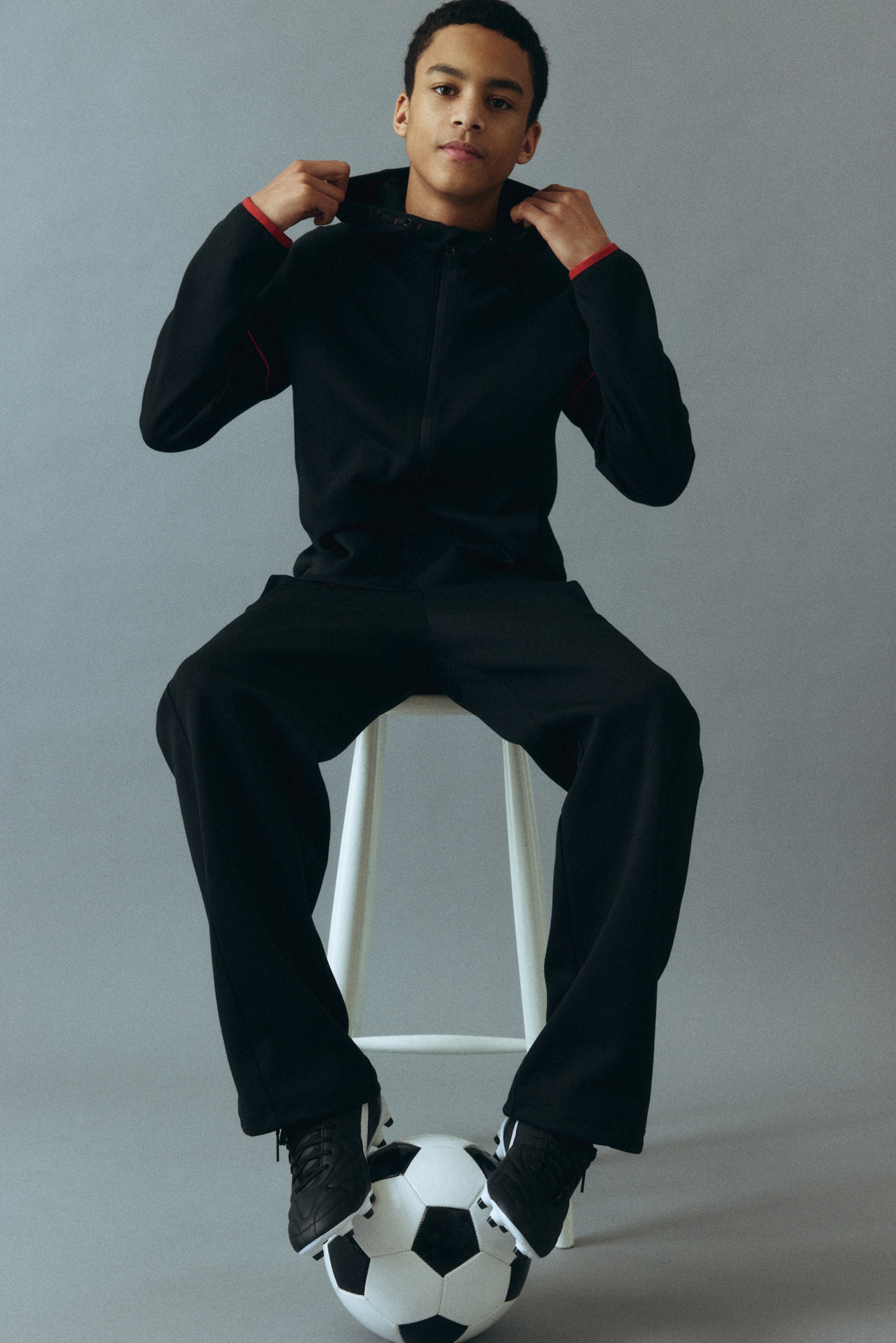 Ver imagem maior: É apresentado um fato de treino preto, composto por um casaco com capuz cujo piping vermelho é visível no interior do capuz e delineia os ombros e as mangas. O casaco apresenta fecho de correr frontal integral, e as calças pretas a condizer têm um corte relaxado.