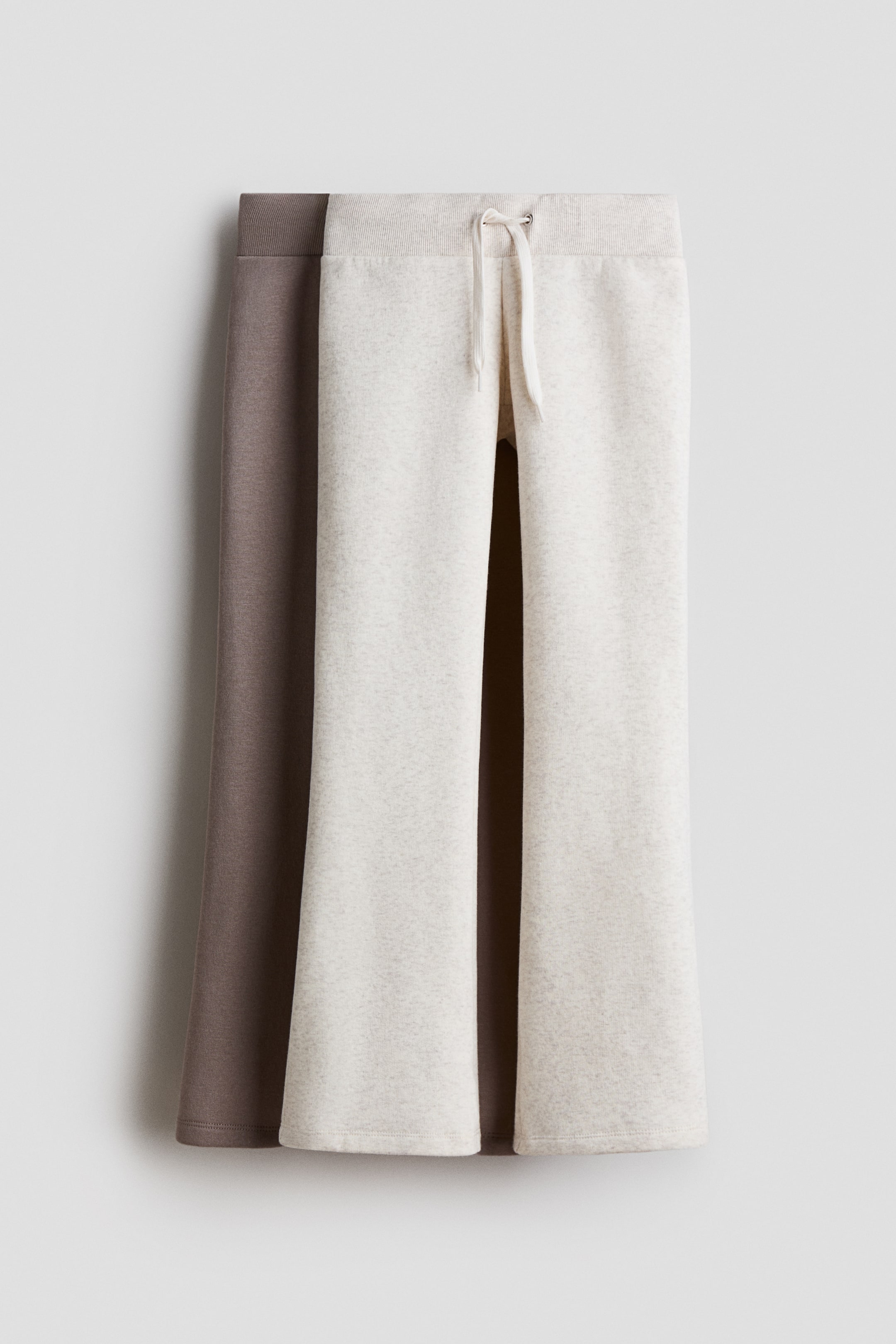 View larger image: Joggers, made from sweatshirt fabric, are featuring an elasticated, drawstring waist and flared legs. The front light beige pair is showing a ribbed elastic waistband with off-white drawstrings, while a dark brown pair is partially visible behind it on the left.