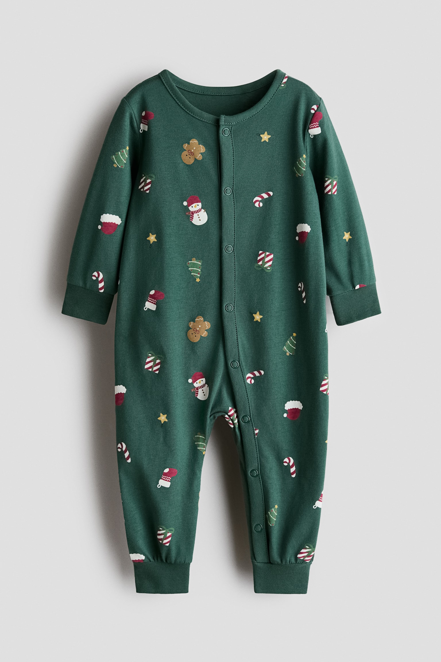 A green cotton-jersey sleepsuit is shown from the front with an all-over festive print of snowmen, gingerbread men, candy canes, and Christmas trees. It features long sleeves with ribbed cuffs, long legs with ribbed hems, and a snap-button closure that extends down the center front and along one leg.
