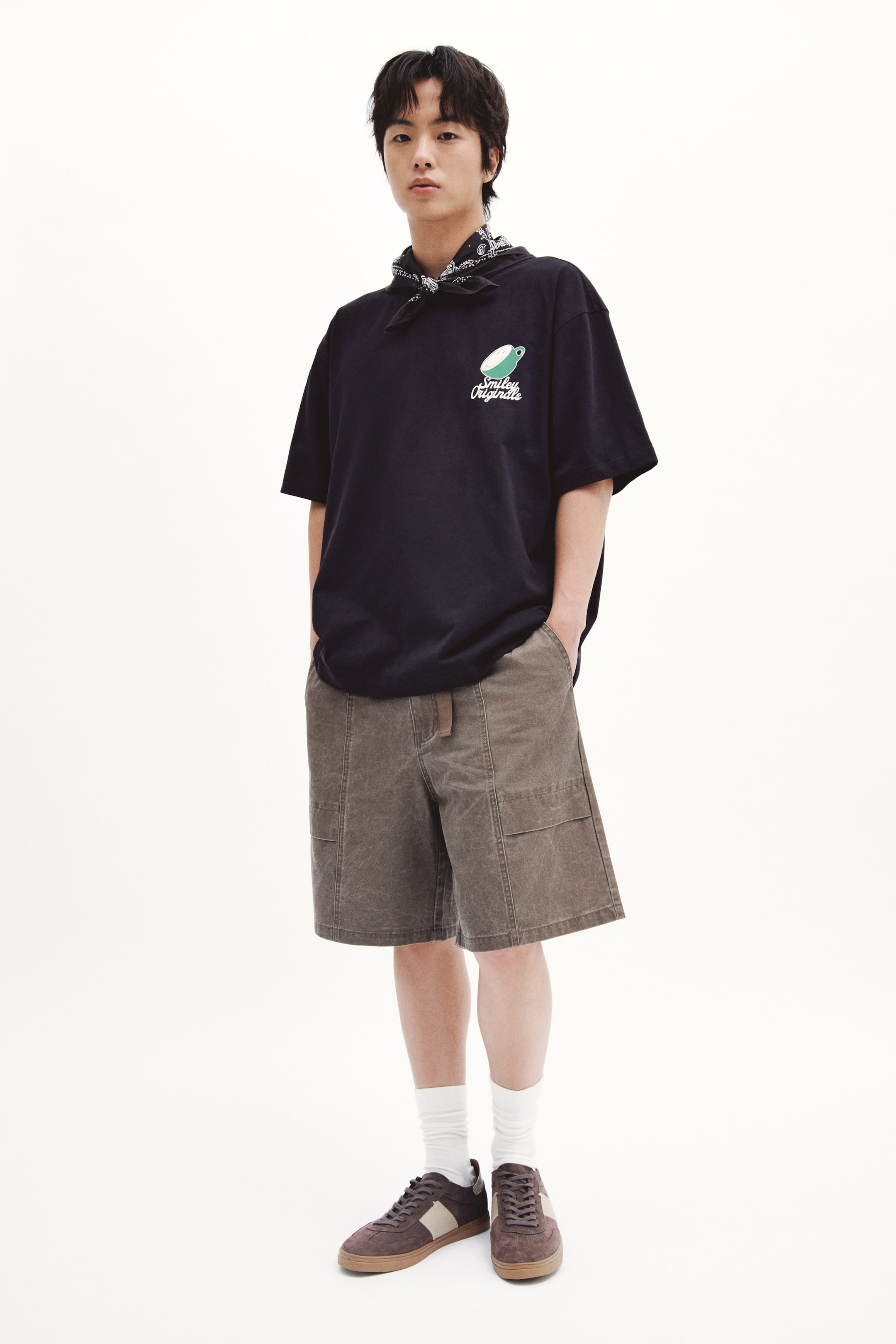View larger image: A black T-shirt featuring a green cup graphic and "Smiley Originals" text is being worn by the model. This T-shirt has a round, rib-trimmed neckline, dropped shoulders, and a loose fit. A patterned bandana is tied around the model's neck, resting against the T-shirt's neckline.