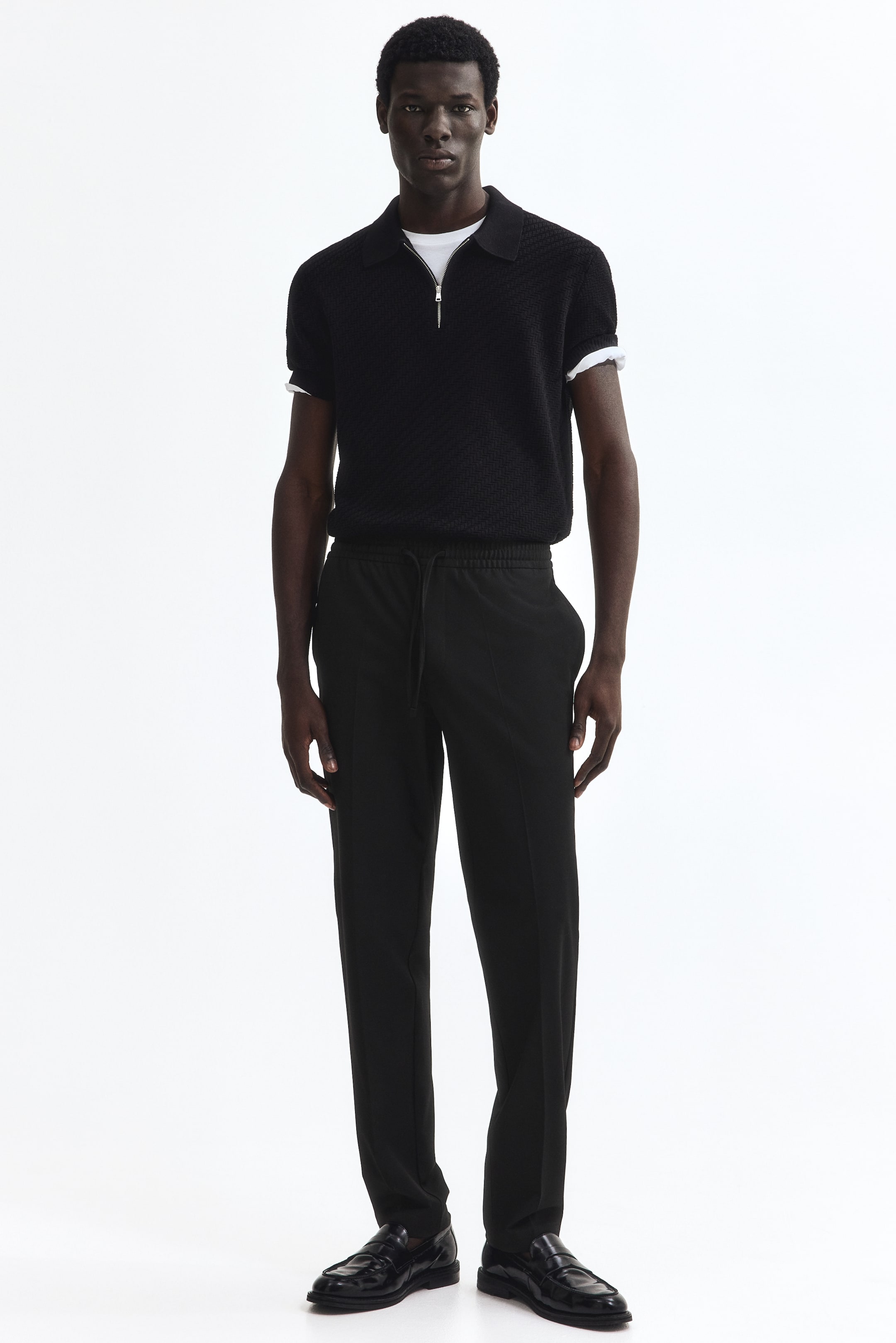 View larger image: A black short-sleeve knit polo shirt with a half-zip closure and textured fabric is layered over a white t-shirt, shown from a front-facing angle. It's paired with black trousers featuring an elastic drawstring waistband and black polished leather loafers.