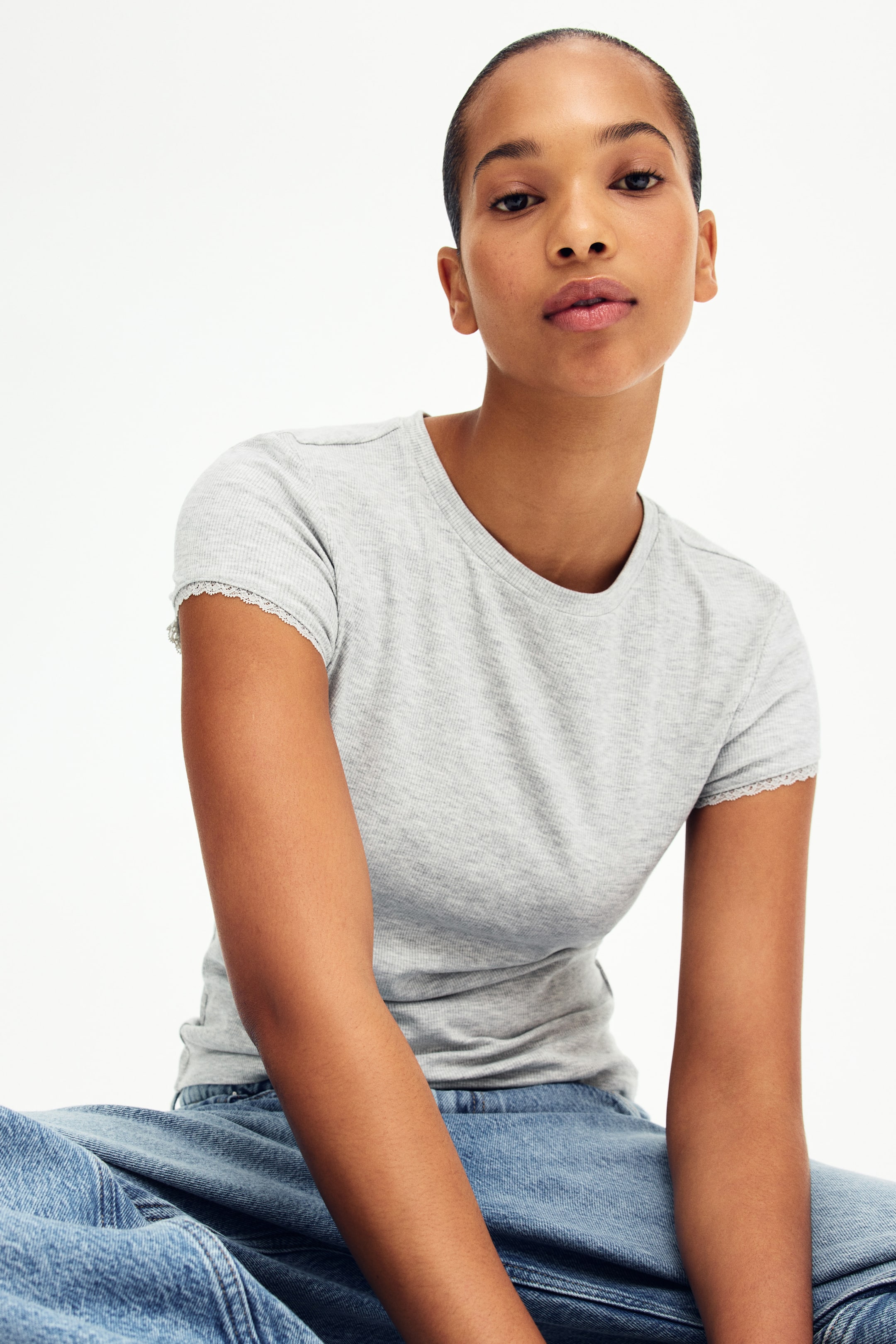 Xem ảnh lớn hơn: Một chiếc áo thun xám nhạt có gân đang được mặc, tạo dáng ôm vừa vặn với cổ tròn và tay ngắn. Viền tay áo được trang trí bằng ren trắng tinh tế.