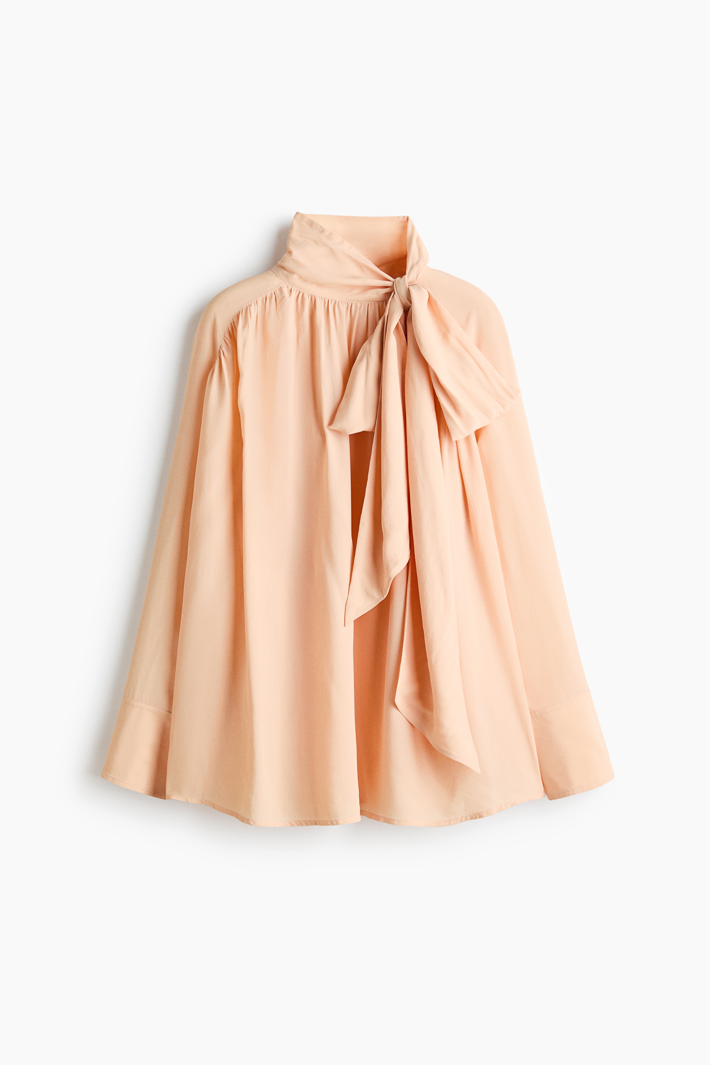 A peach-coloured, relaxed-fit blouse in a lightweight fabric is worn by the model, featuring a high collar with long, draped neck ties and gathered raglan sleeves. The model is posing with one hand resting on their hip, styling the blouse with light-wash, wide-leg blue jeans.