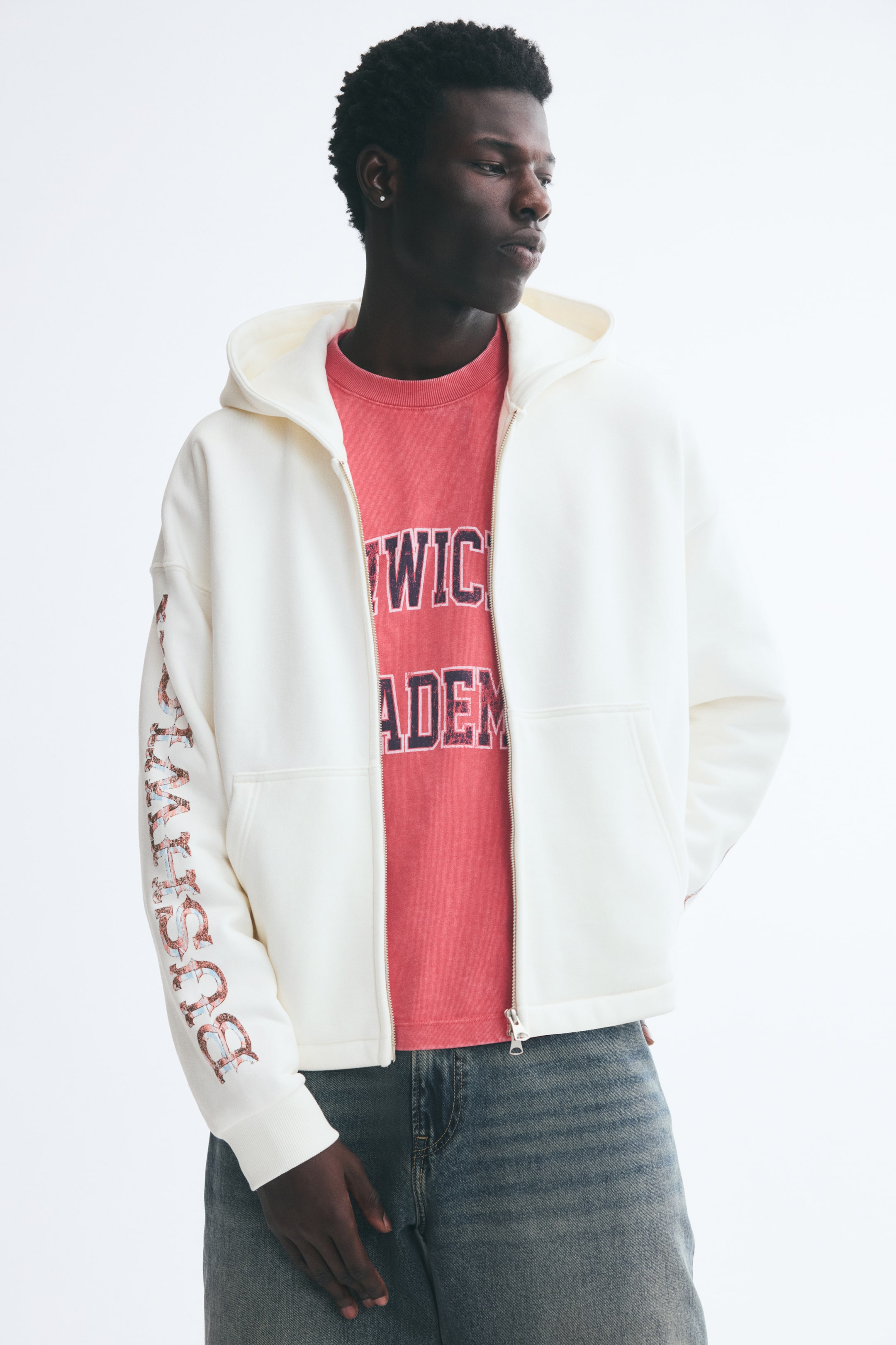 View larger image: An off-white zip-up hoodie with a graphic print on the right sleeve is being worn by the model. The hoodie is styled open, layered over a coral-red graphic T-shirt and blue denim jeans.