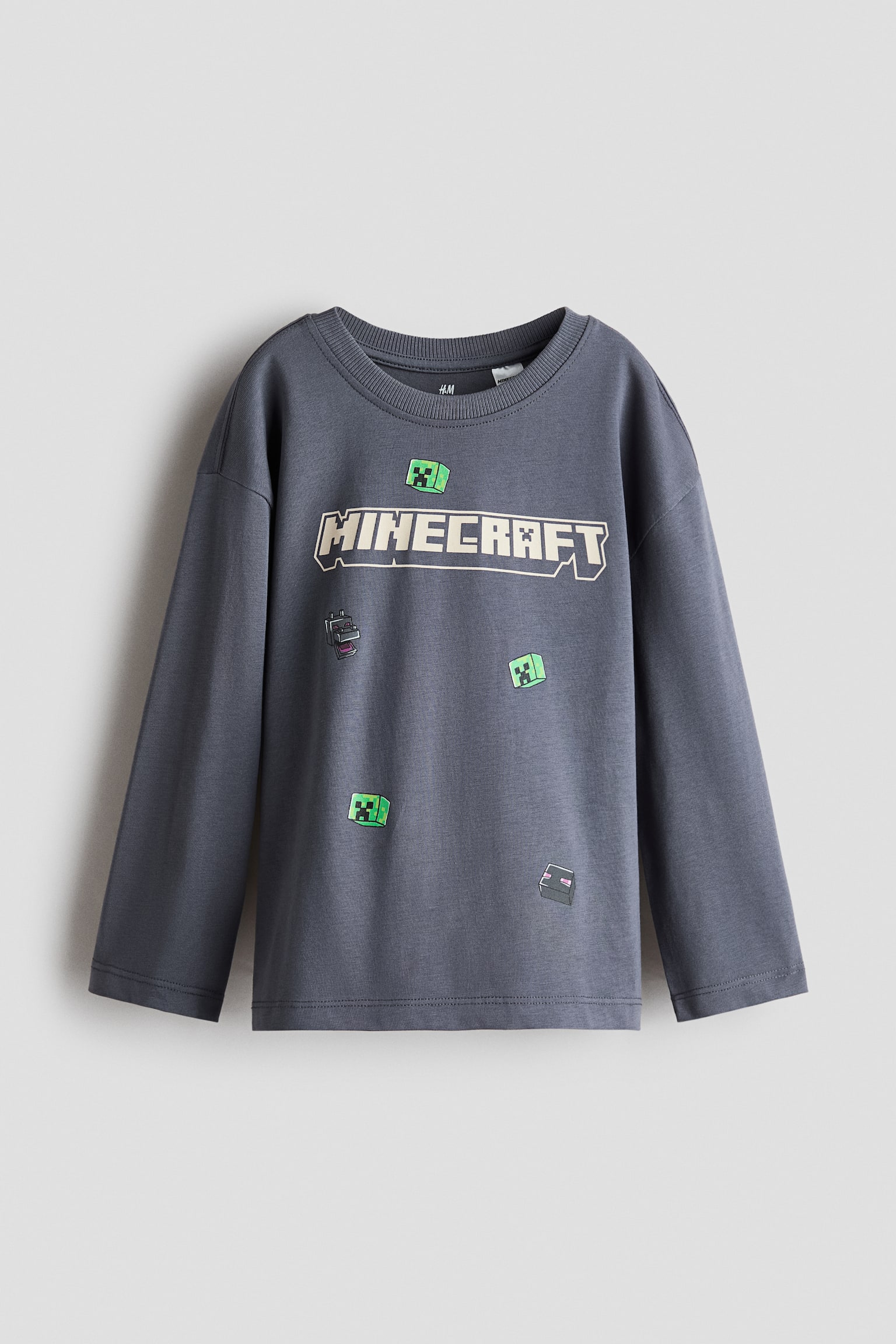 A dark grey, long-sleeved tee is shown from the front, featuring a round, rib-trimmed neckline, dropped shoulders, and a straight-cut hem. The tee is displaying a printed beige 'MINECRAFT' logo and pixelated game characters across the chest.